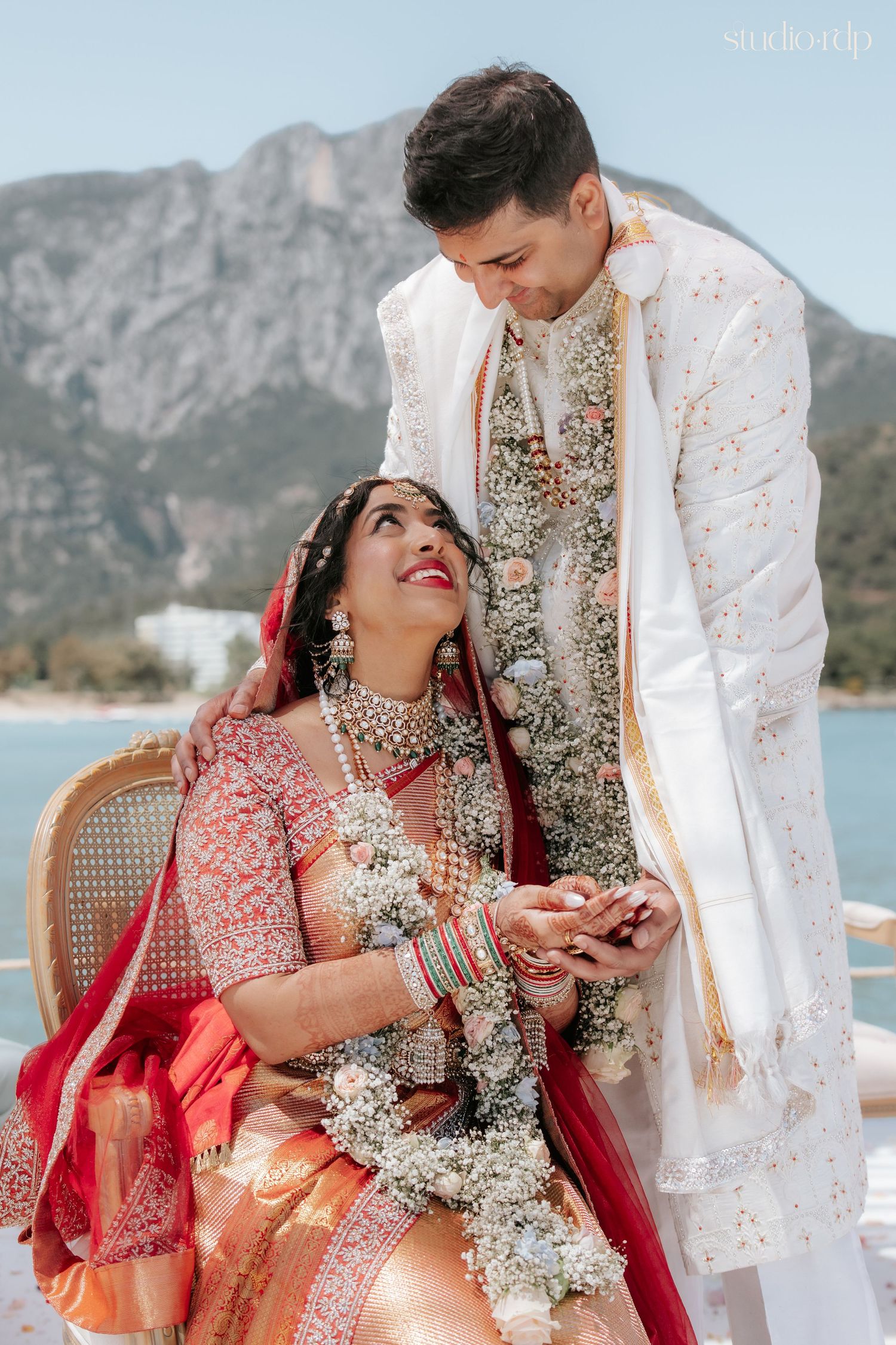 Indian bride in red lehenga and groom in white sherwani share a joyful moment at outdoor wedding ceremony.