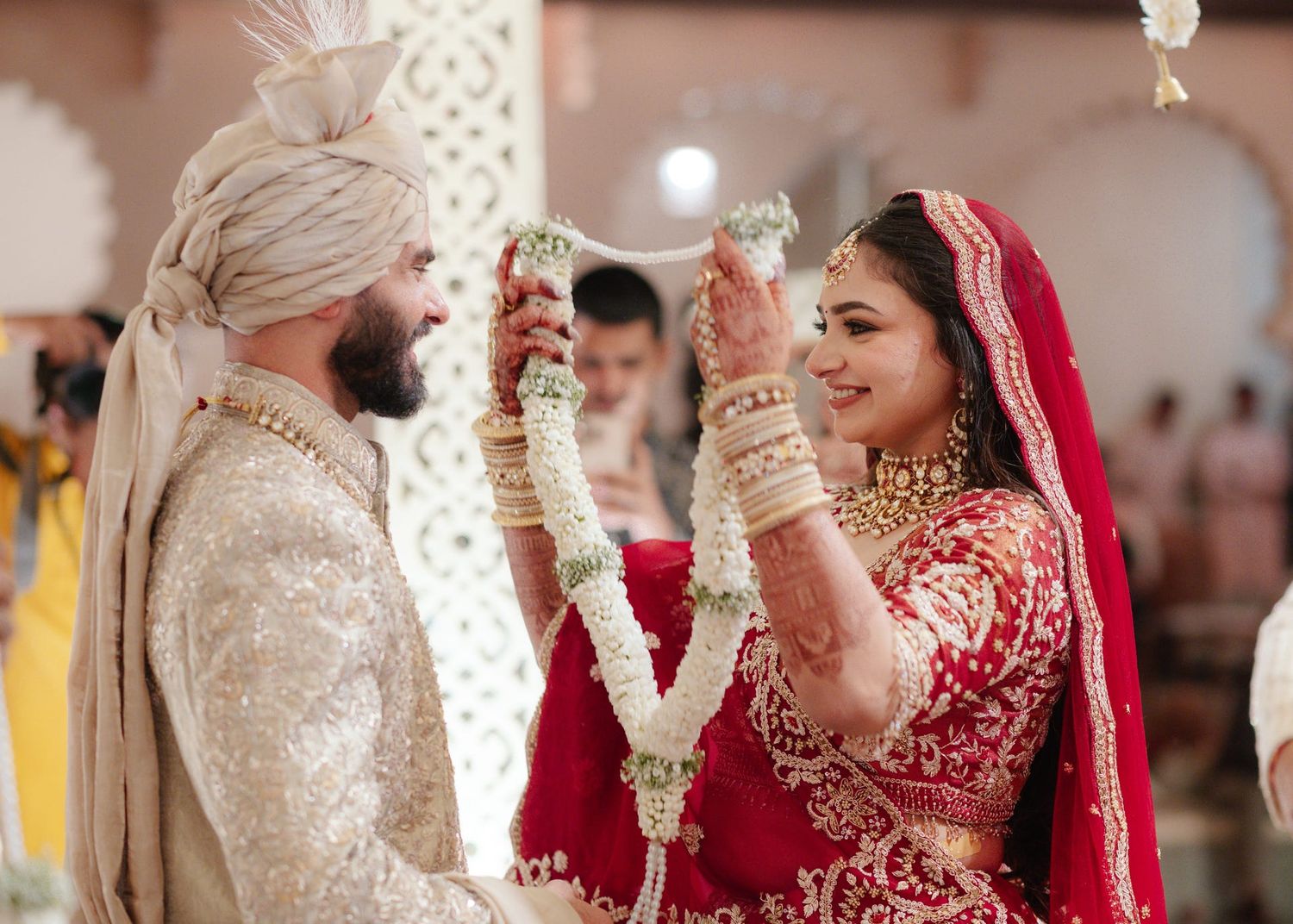 Indian bride in red lehenga and groom in ivory sherwani exchange garlands during traditional Hindu wedding ceremony.