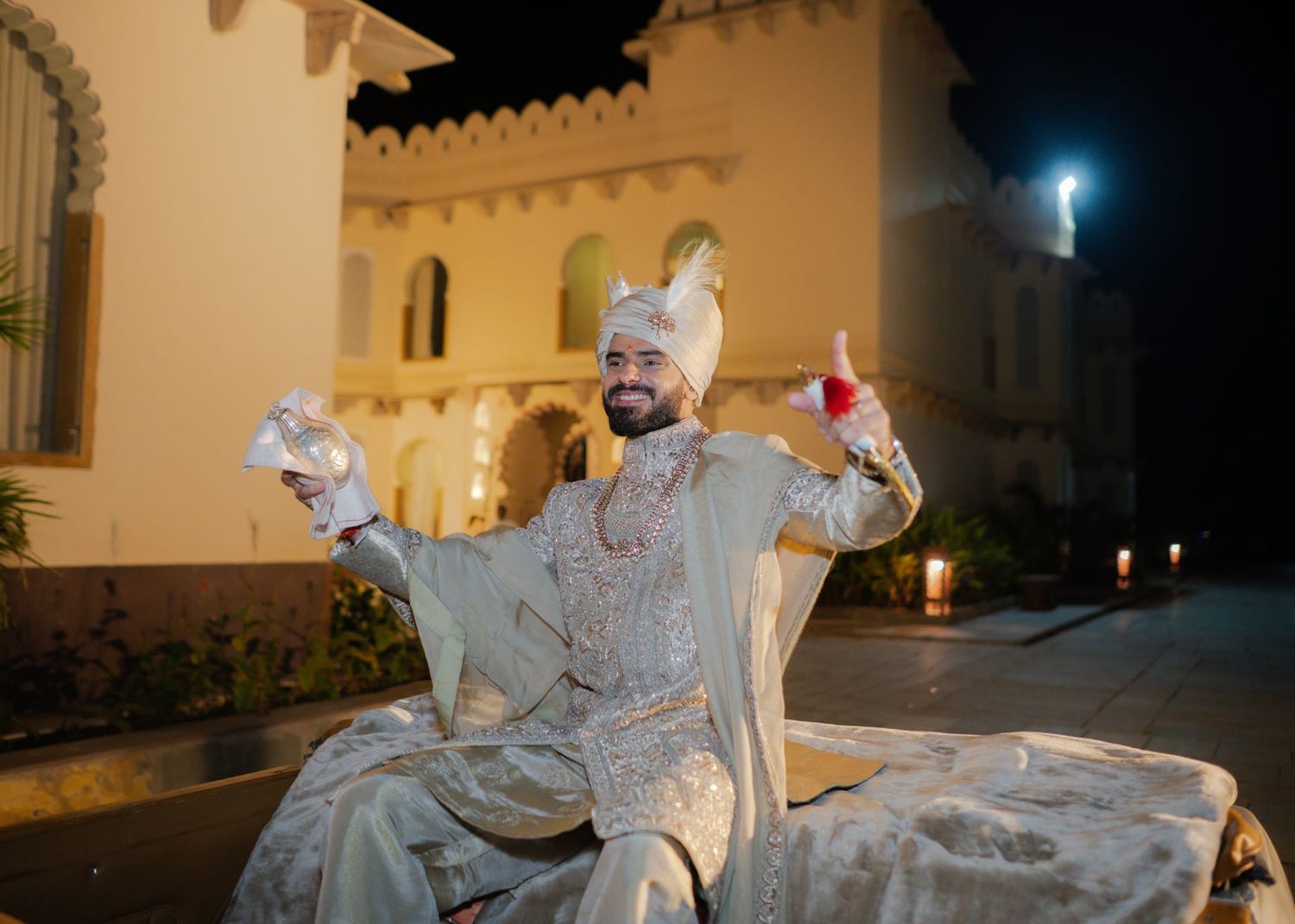 Indian groom in ivory sherwani and turban poses joyfully outdoors at night during wedding celebration.