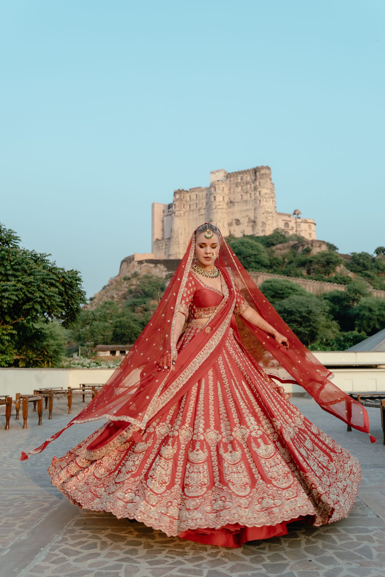 Ornate red wedding lehenga photographed in spinning motion against historic fortress backdrop.