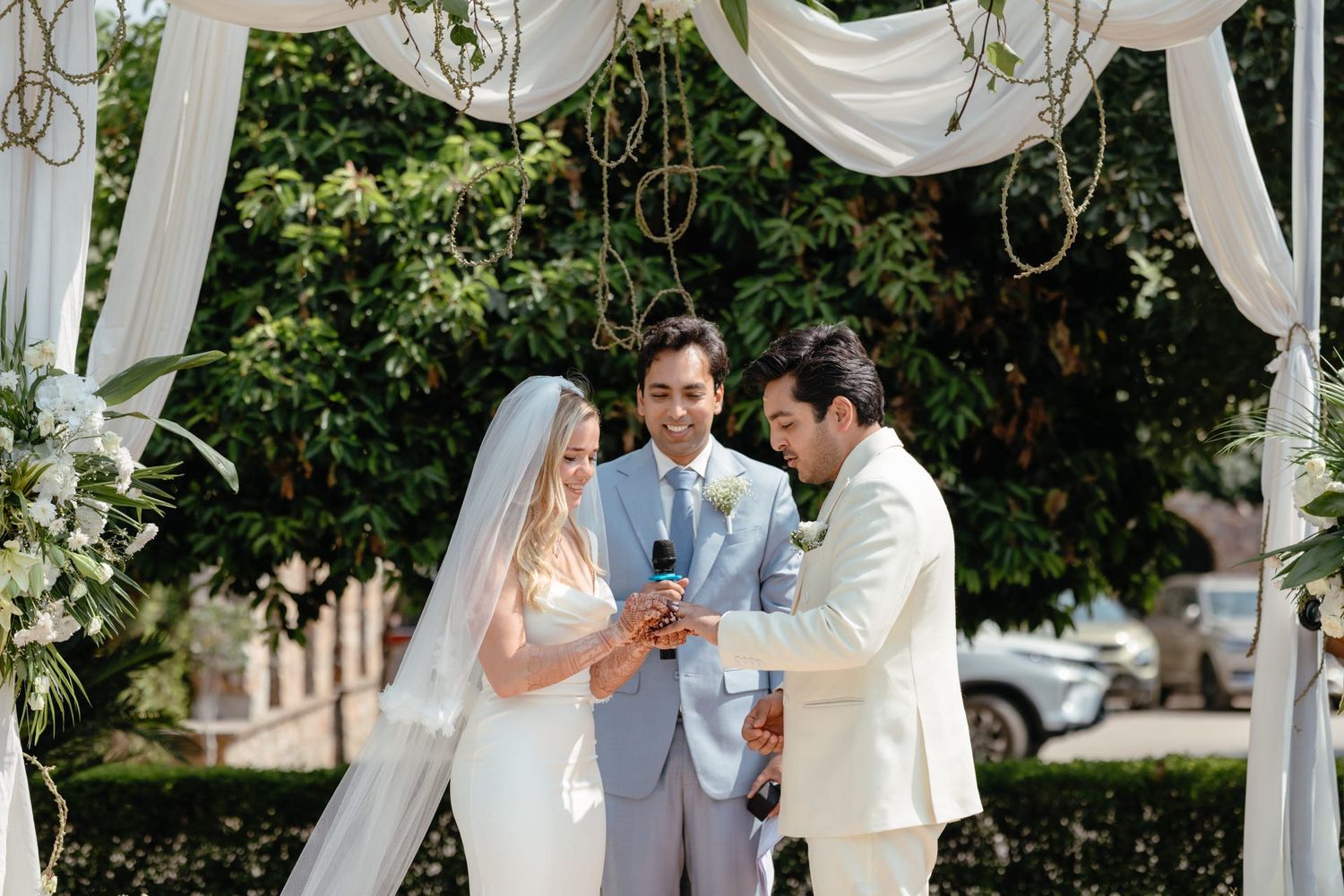 An outdoor wedding ceremony with an officiant in gray and couple in white beneath a decorated arch.