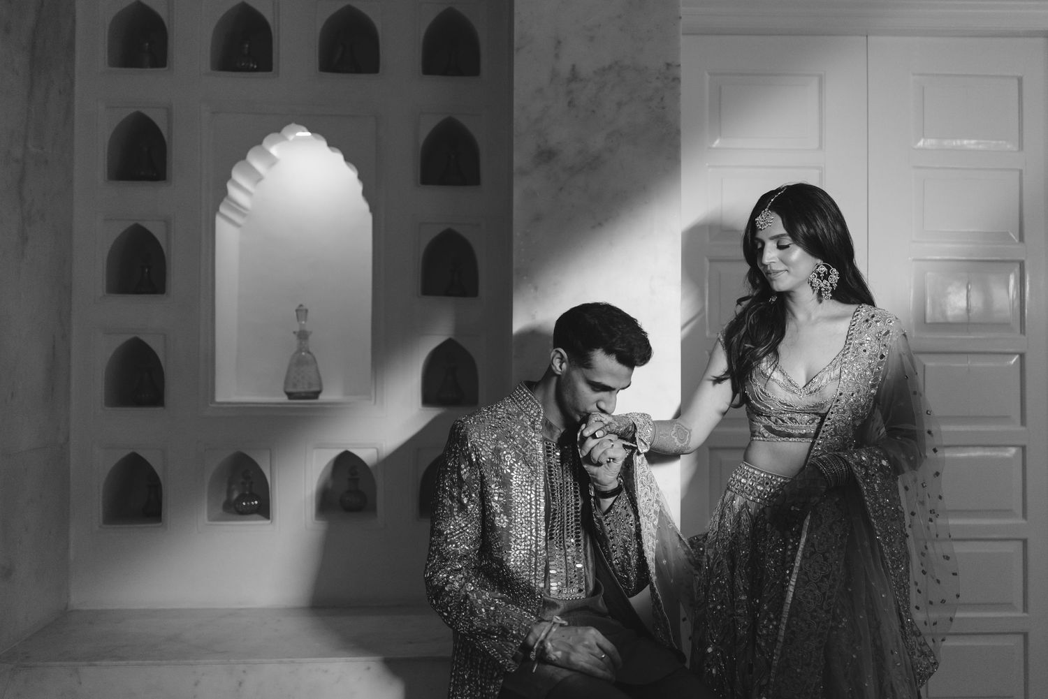 A romantic black and white portrait of a couple in ornate Indian wedding attire near arched windows.