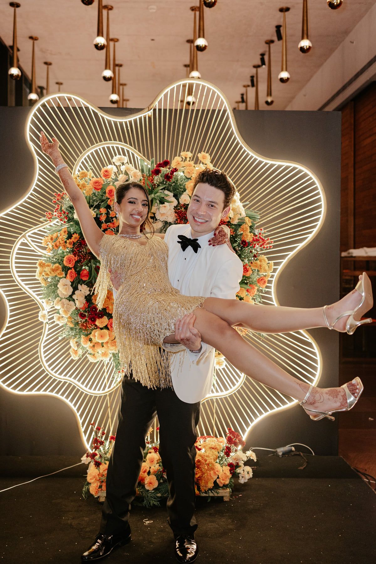 A couple poses in front of an illuminated wing-shaped light installation with floral decorations.