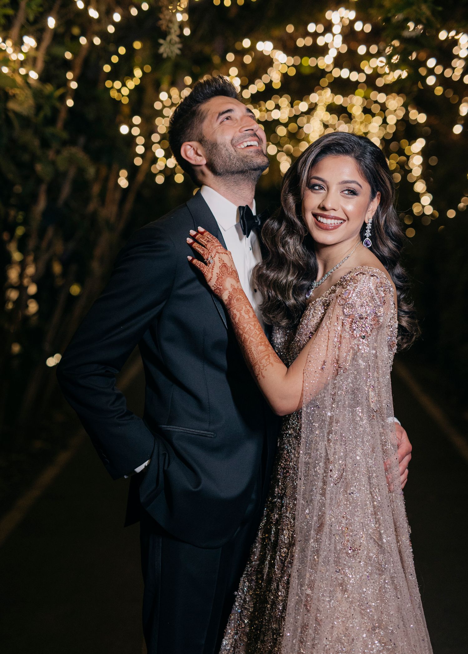 A couple celebrates amid twinkling lights, with her wearing a sparkly beige gown and him in a dark suit.