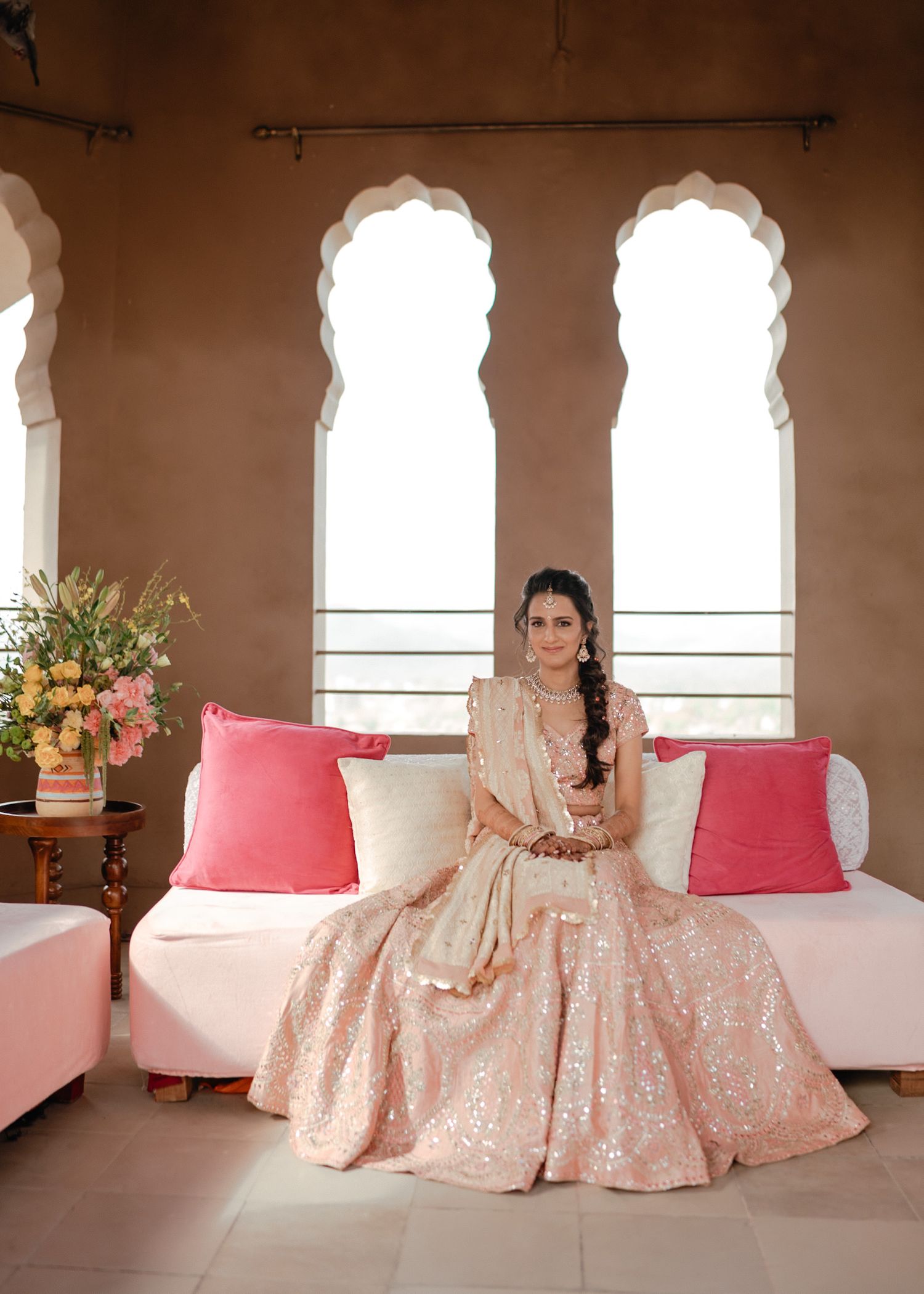 A person wearing a blush pink sequined gown sits elegantly on a white sofa with coral pillows in an arched window setting.