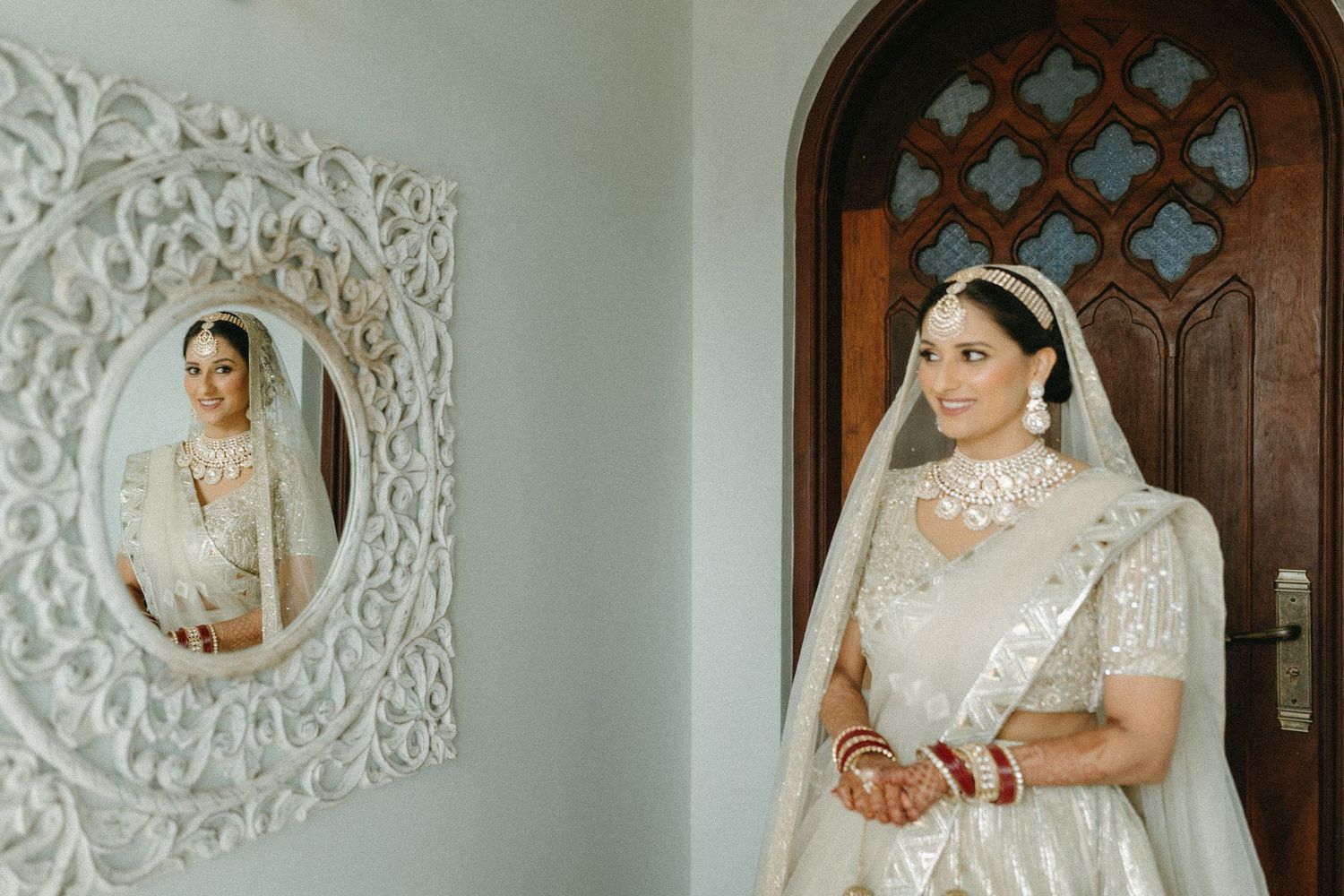 Traditional bride in white Indian wedding attire stands near an ornate wall mirror and wooden door with Gothic details.