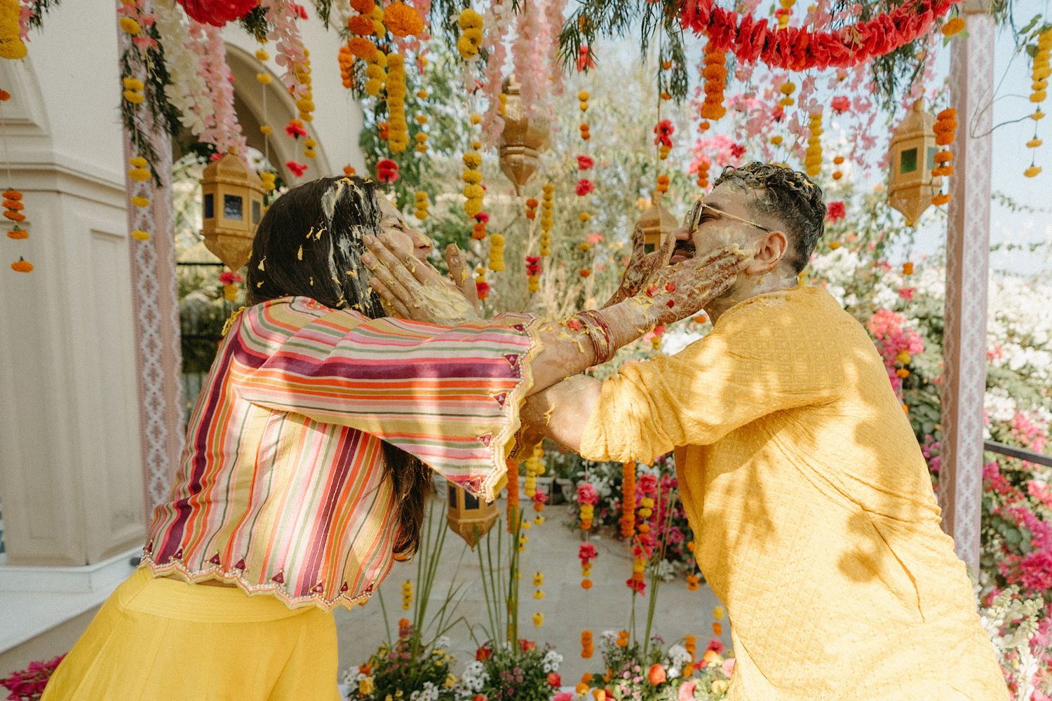 Two people in yellow outfits playfully smear color on each other during a festive celebration with floral decorations.