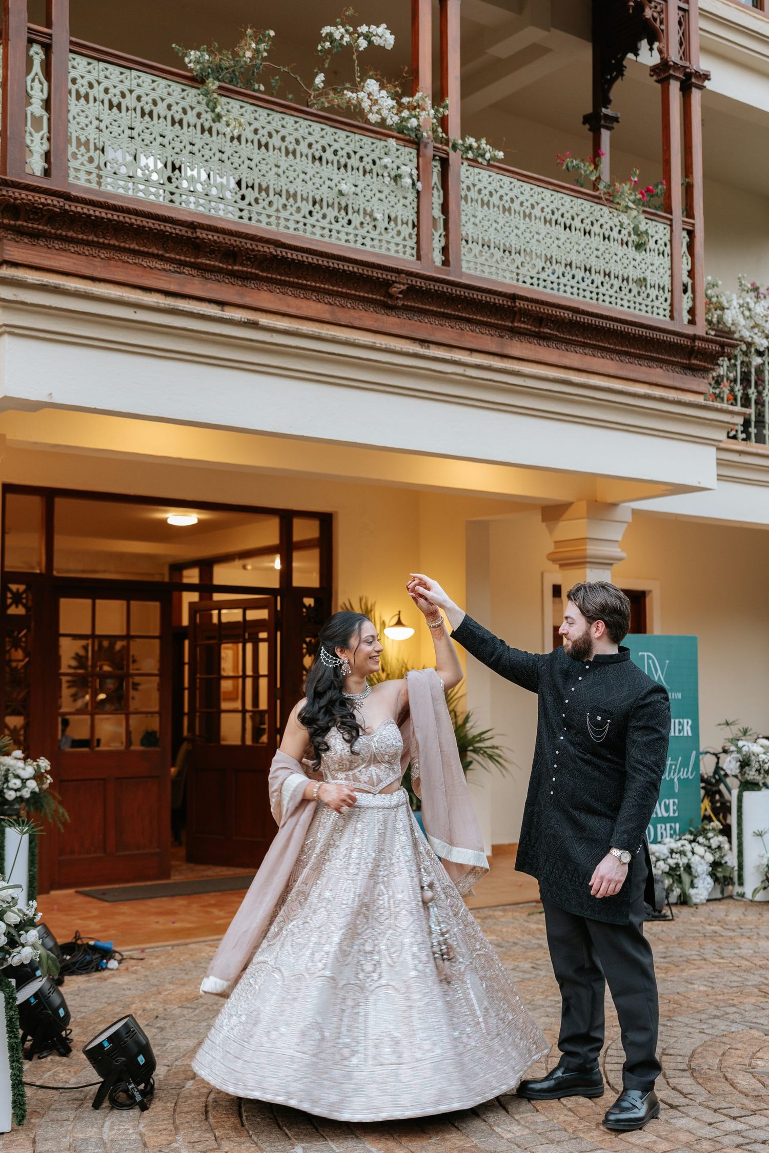 Bride in silver lehenga twirls with groom in black sherwani outside elegant venue with floral balcony.