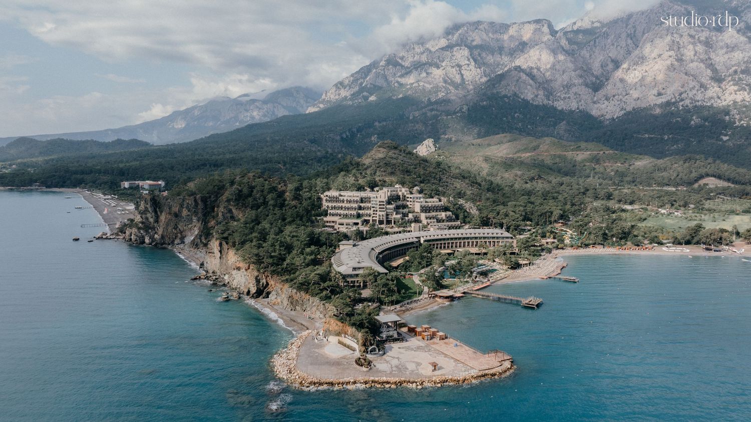 Aerial view of a luxury coastal resort nestled among lush cliffs with turquoise sea and mountain backdrop.