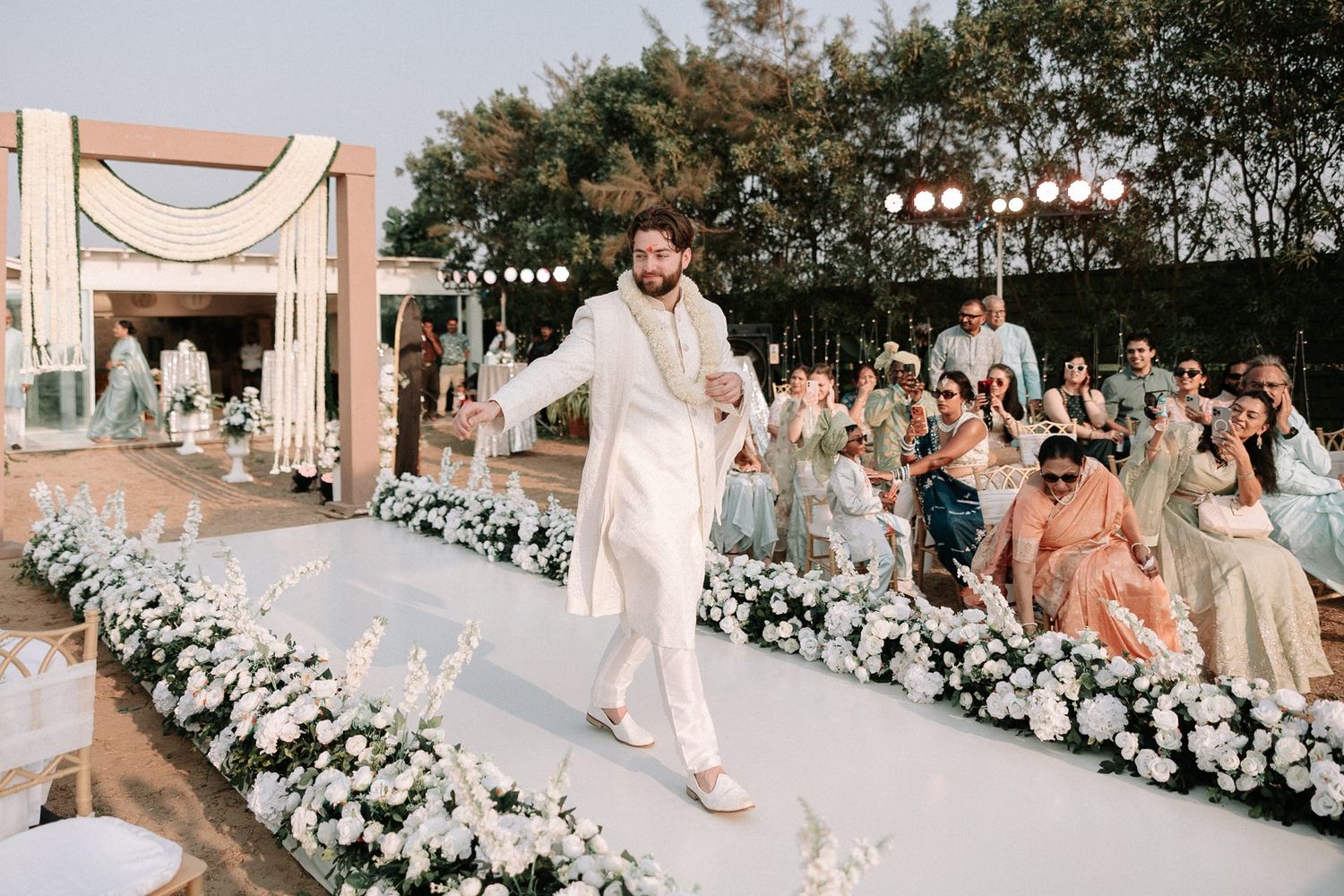 Groom in white sherwani dancing at an outdoor Indian wedding ceremony with floral decorations.