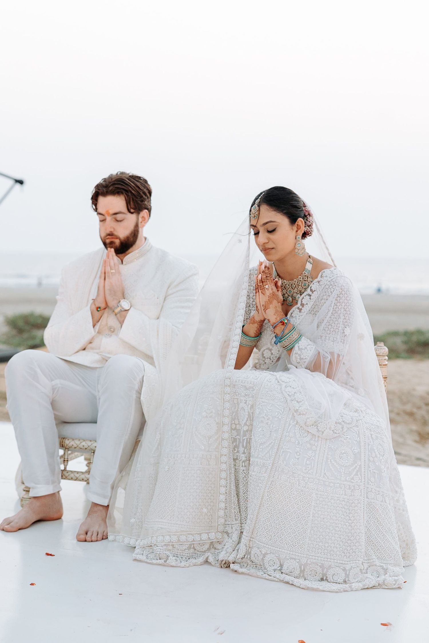 Couple in white traditional Indian wedding attire seated with hands in prayer pose at a beach ceremony.