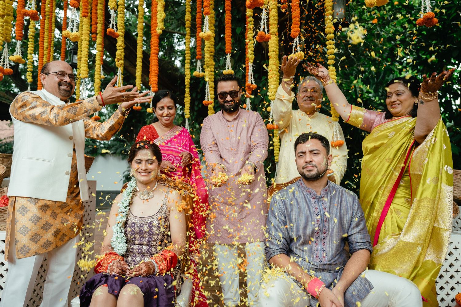 Joyful Indian wedding celebration with family tossing flower petals over bride and groom under marigold decorations.