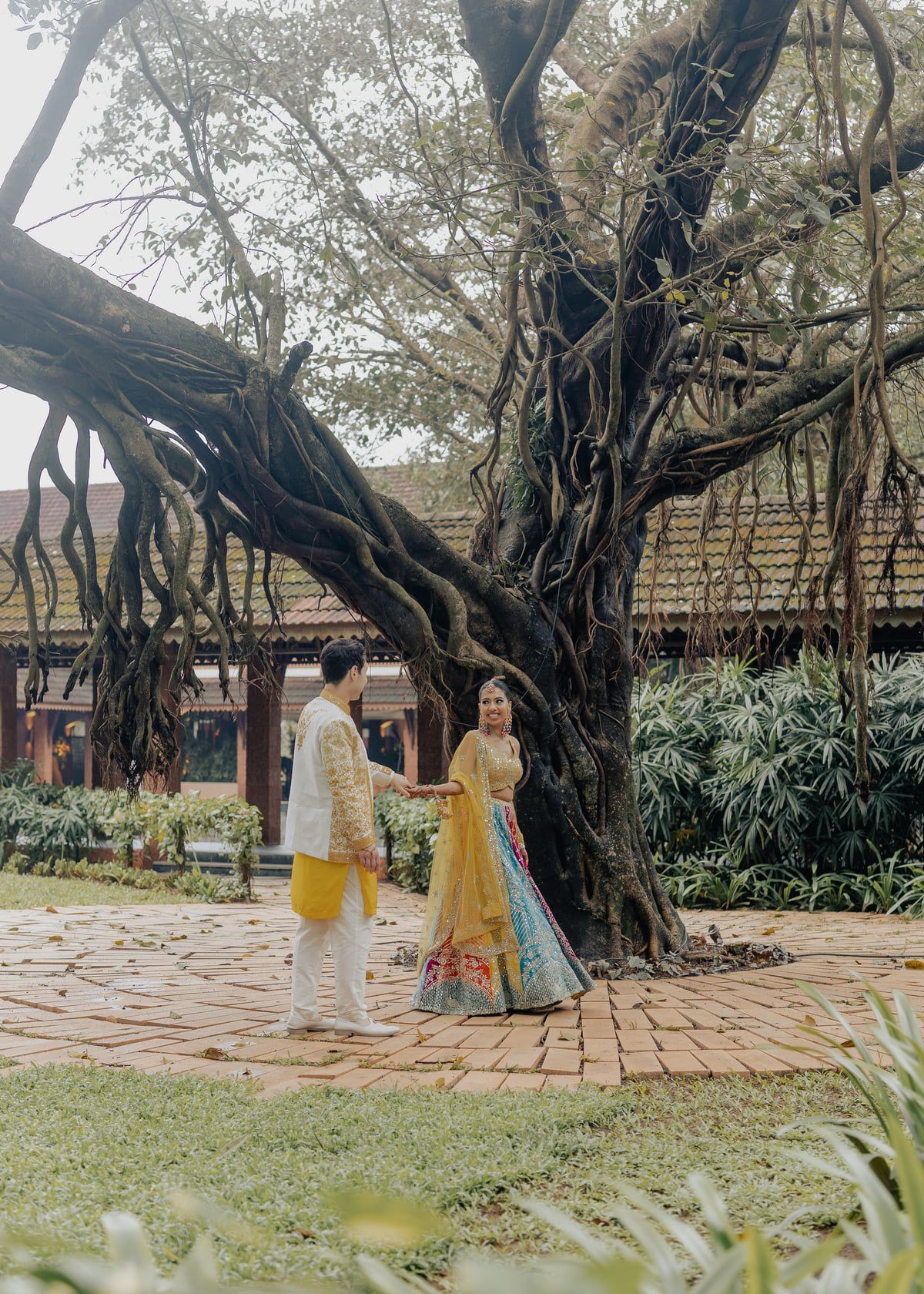 Couple in traditional Indian wedding attire holding hands under a large banyan tree in a lush garden courtyard.