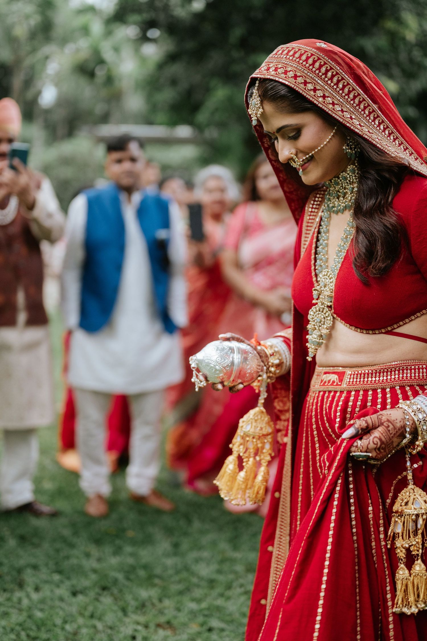 Indian bride in red lehenga with gold jewelry performs wedding ritual outdoors while guests watch and photograph.