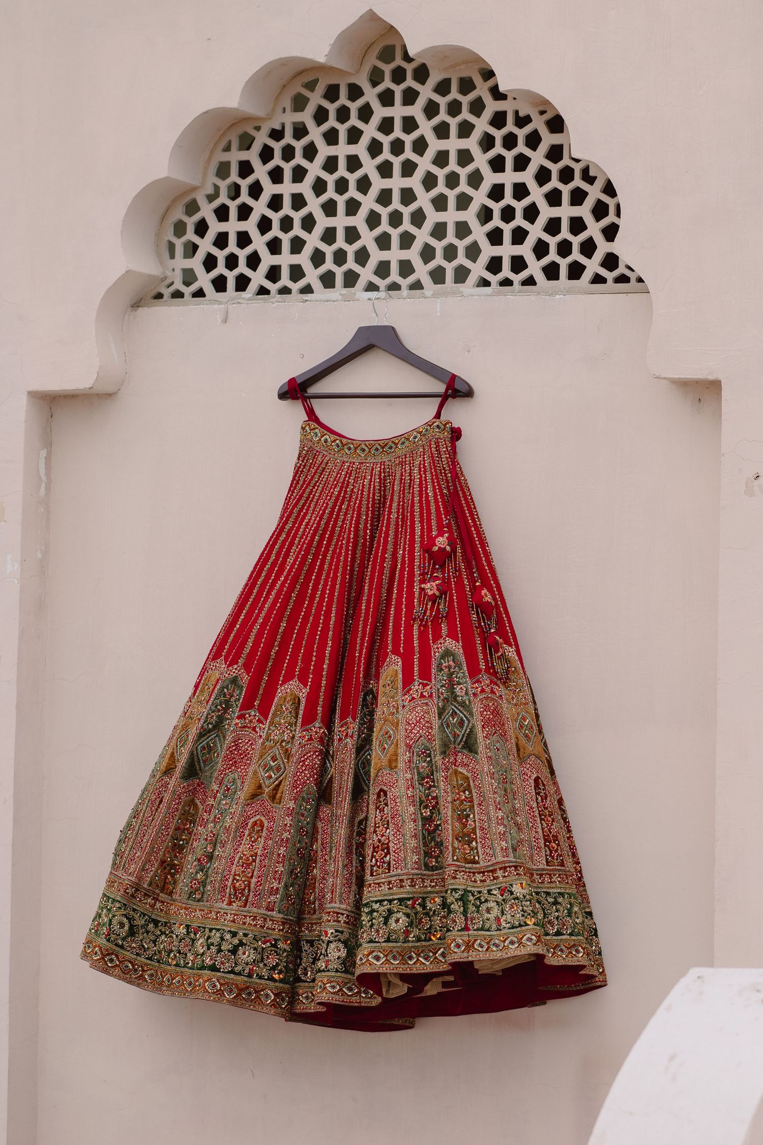 Red bridal lehenga with intricate gold embroidery displayed on a hanger against ornate white Mughal arch.