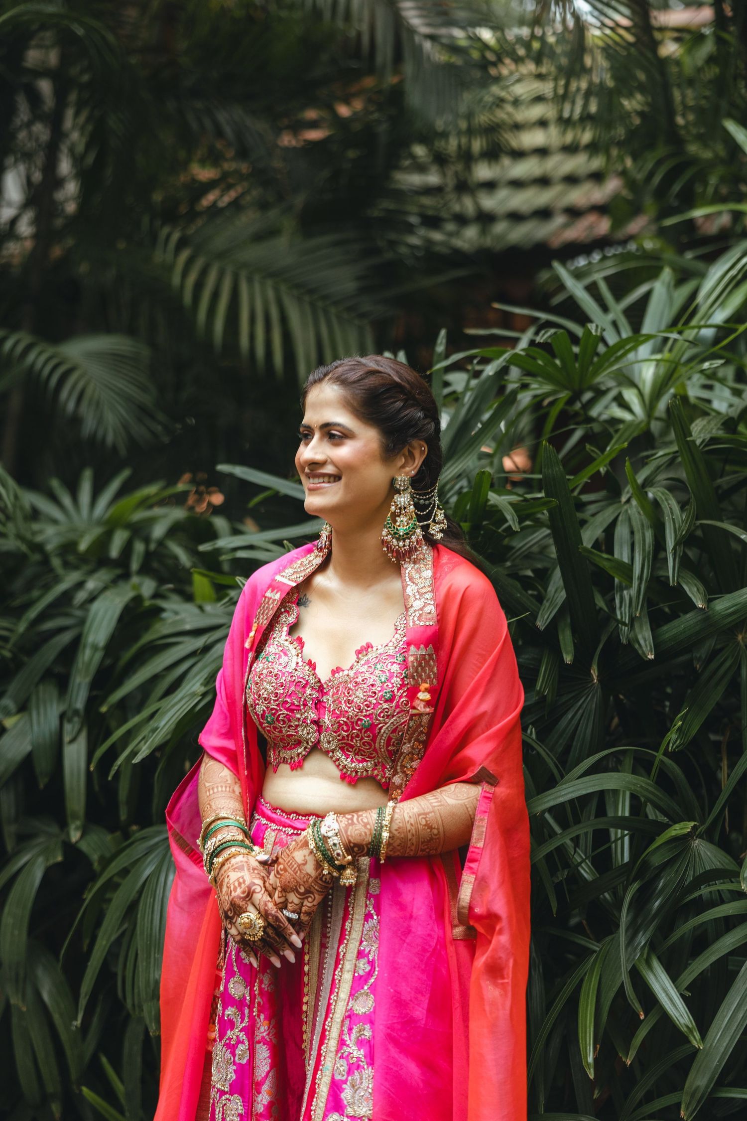 Smiling Indian bride in pink lehenga with mehndi hands, green earrings, standing outdoors among tropical plants.