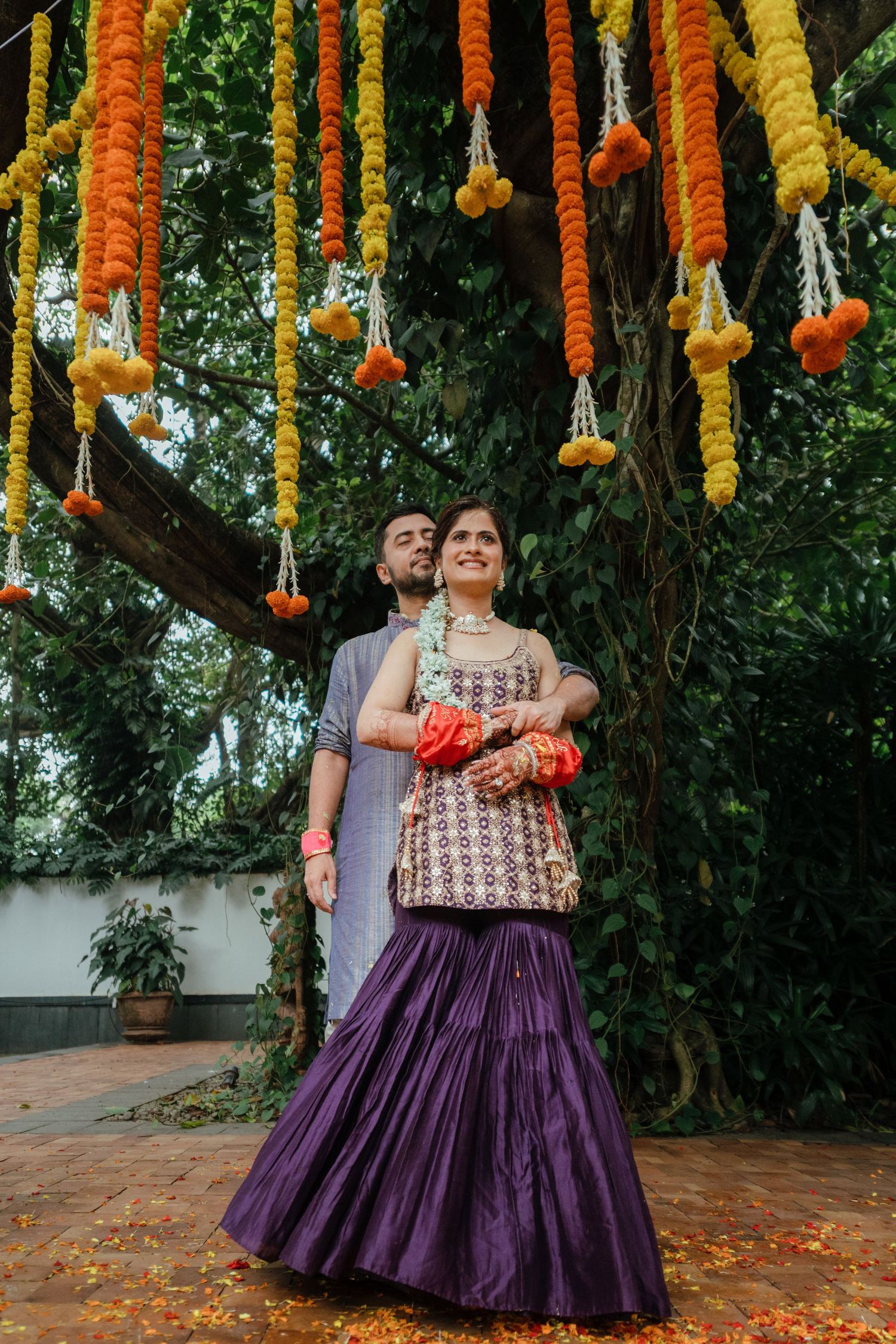 Indian couple in traditional attire pose under colorful marigold garlands outdoors during a festive wedding celebration.
