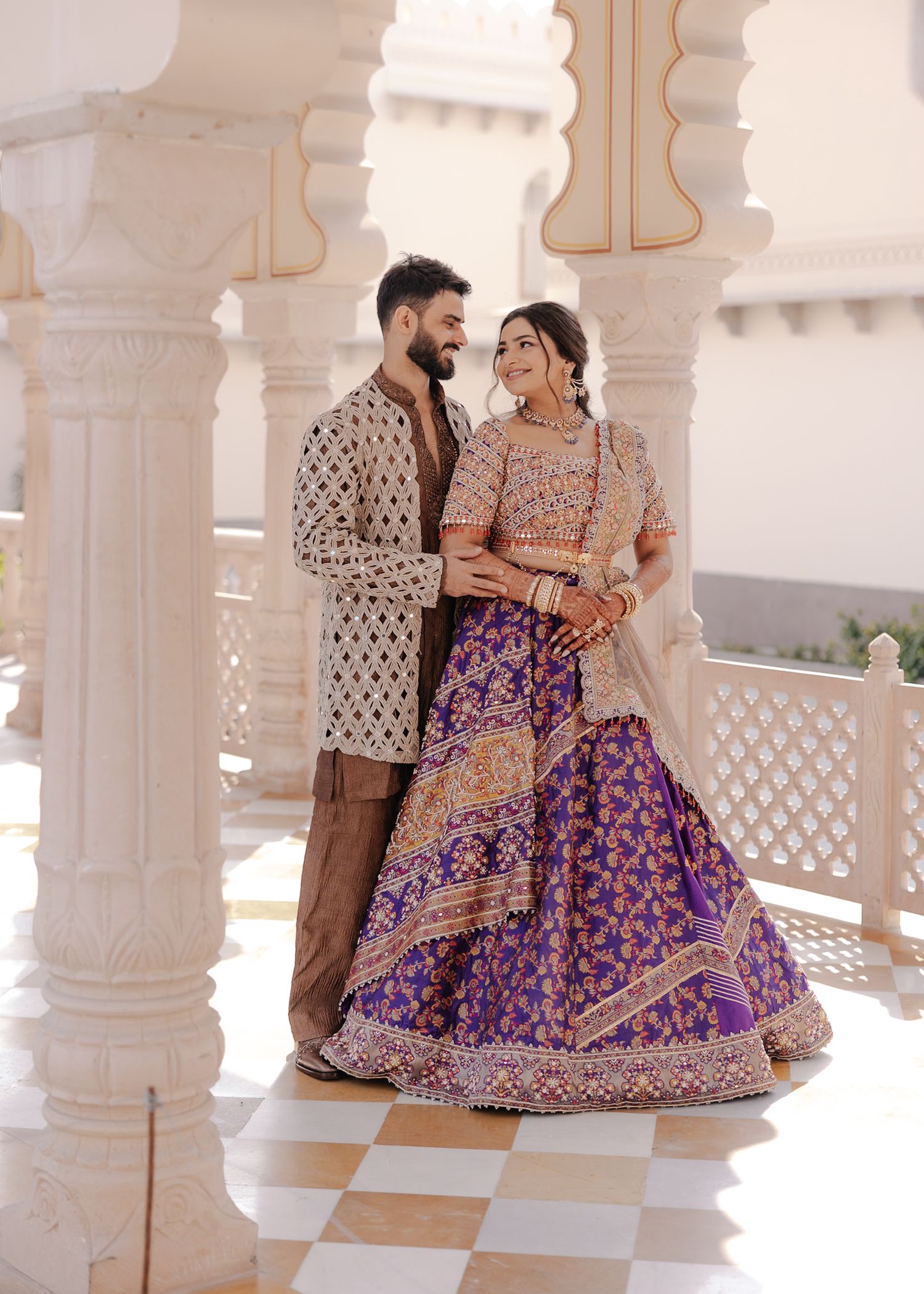 Couple in traditional Indian wedding attire posing between ornate white marble columns at a grand venue.