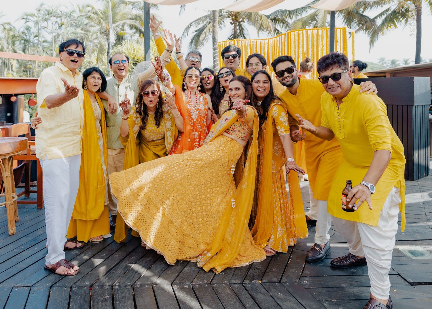 Wedding guests strike cheerful poses in coordinated yellow outfits during celebration at outdoor venue near palm trees.