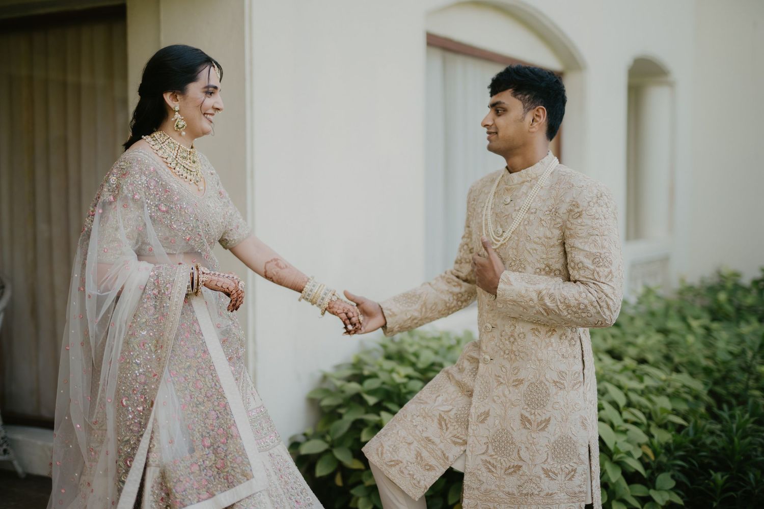 A couple in traditional beige and cream South Asian wedding attire hold hands while smiling at each other outdoors.