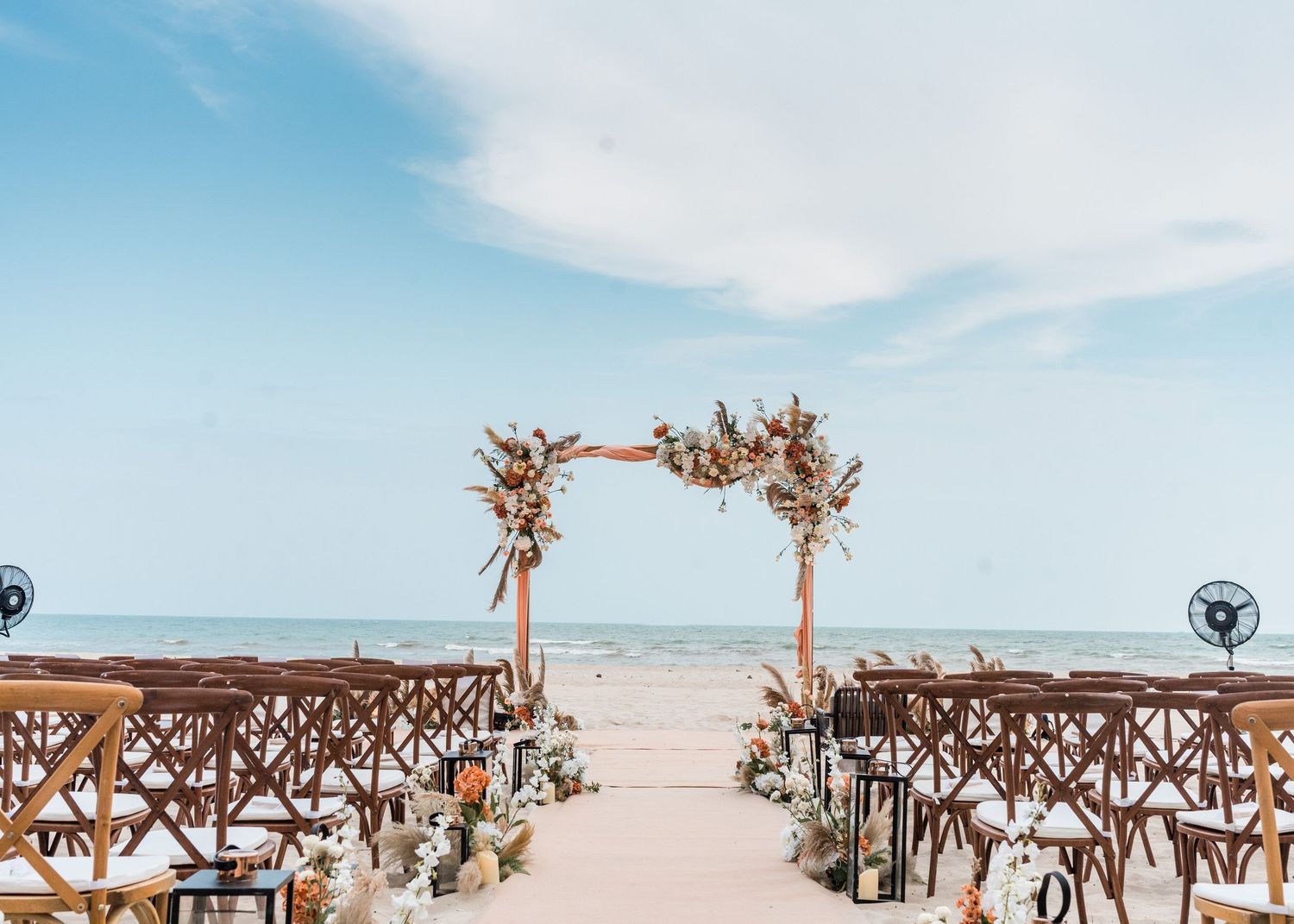 Rustic wooden wedding arch with dried flowers stands on a sandy beach with cross-back chairs and ocean views.