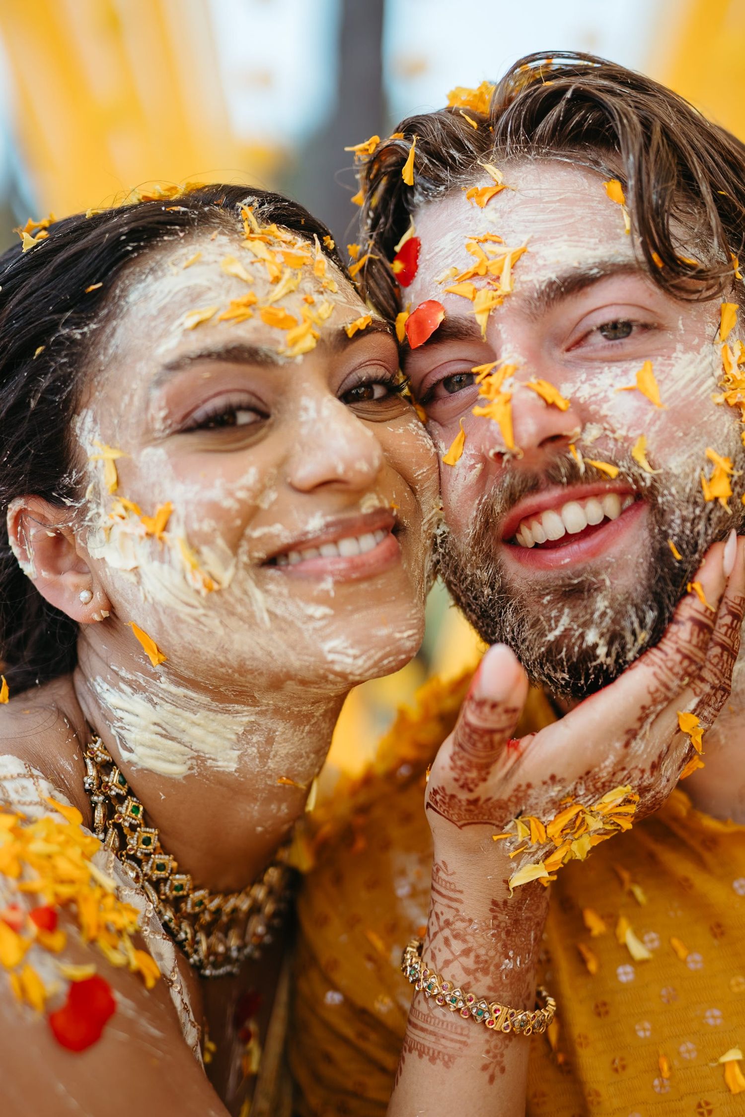Joyful couple covered in haldi paste and marigold petals smiling during Indian wedding haldi ceremony.