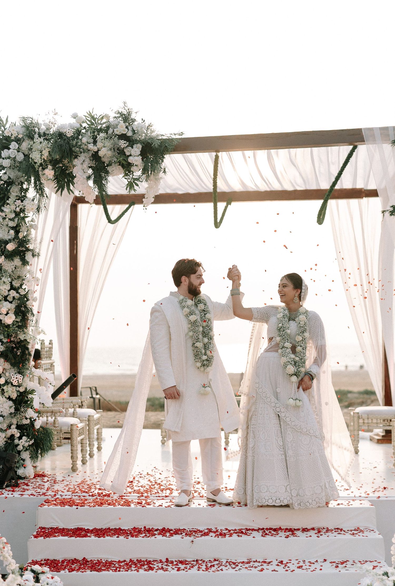 Bride and groom in white traditional attire holding hands under floral mandap with rose petals at beach wedding.