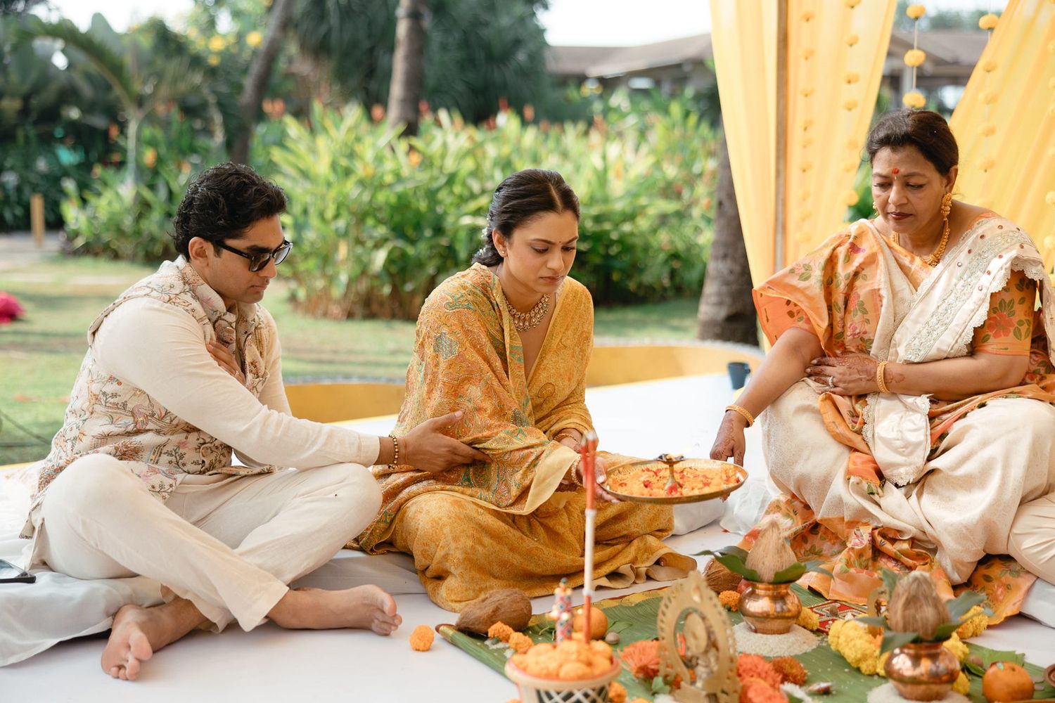 Three people in traditional Indian attire performing a Hindu puja ceremony with flowers and offerings outdoors.