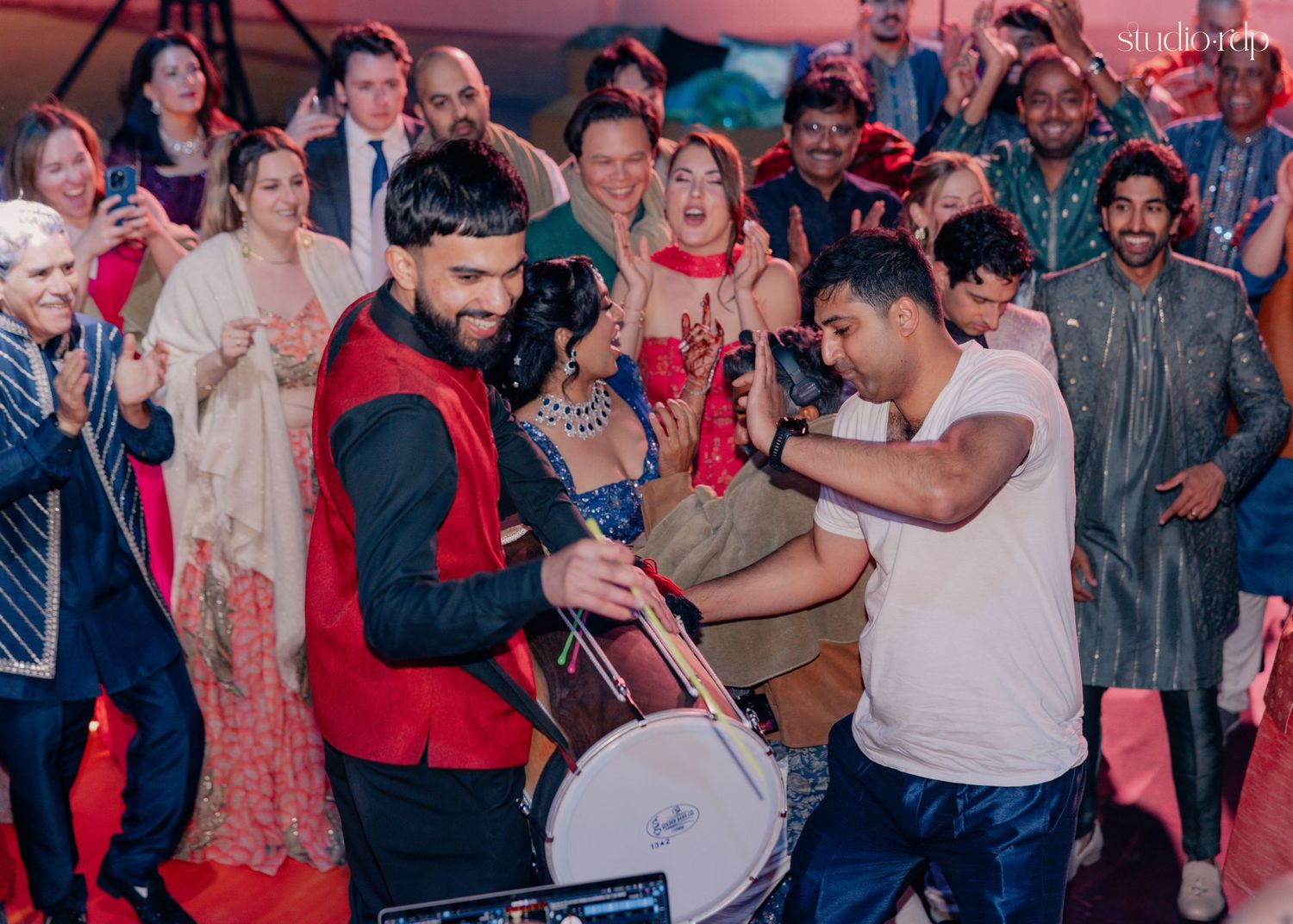Guests celebrate at a vibrant South Asian wedding reception as a man plays a dhol drum on the dance floor.