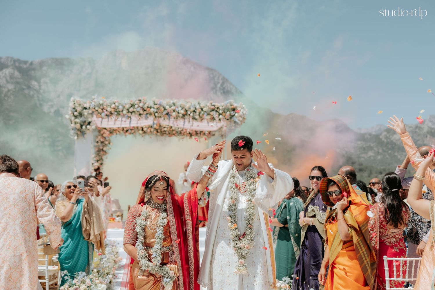 Newlywed Indian couple exits ceremony amid colorful powder and confetti surrounded by joyful guests outdoors.