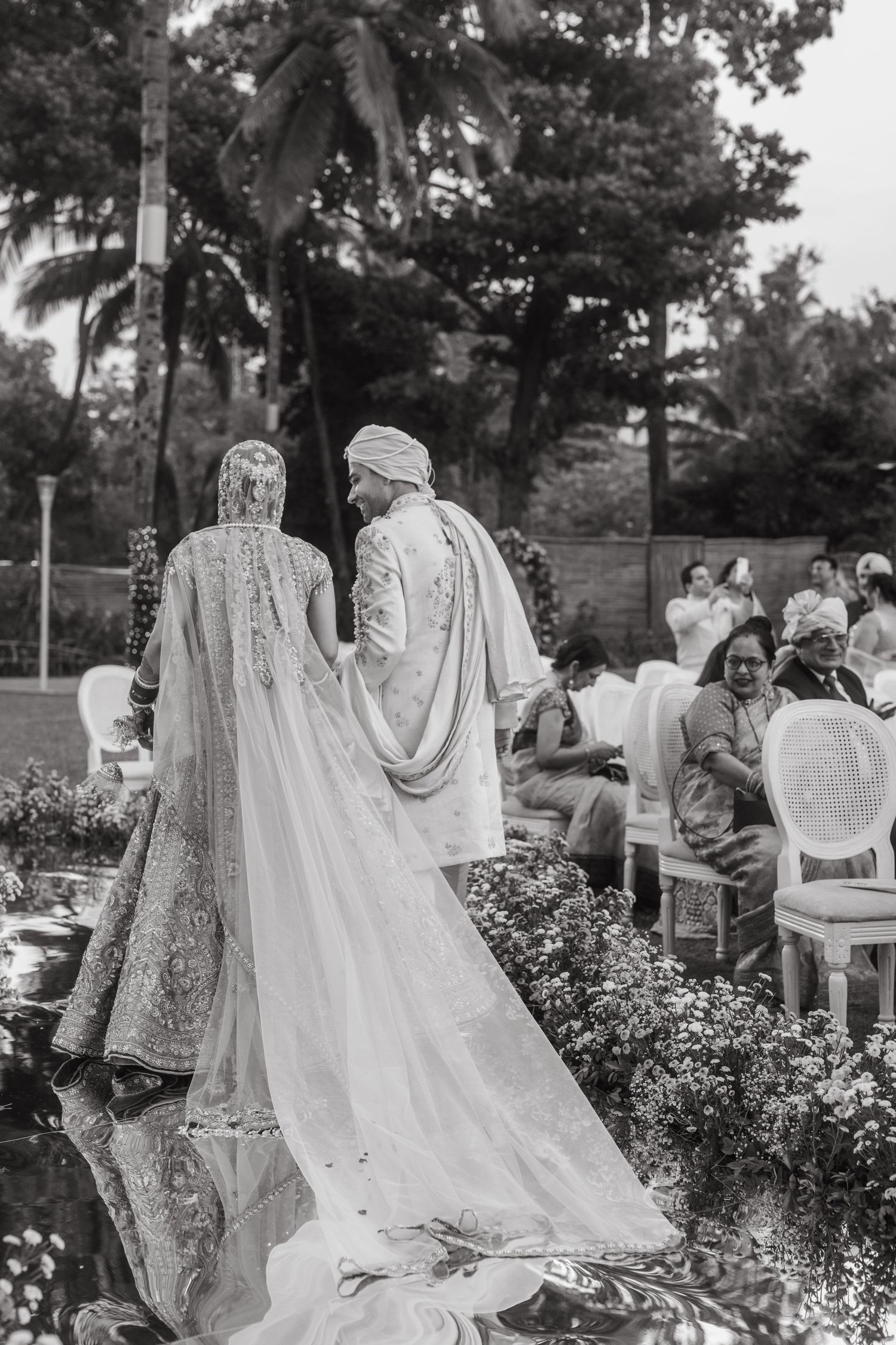 A black and white photograph of a couple in traditional wedding attire during an outdoor garden ceremony.