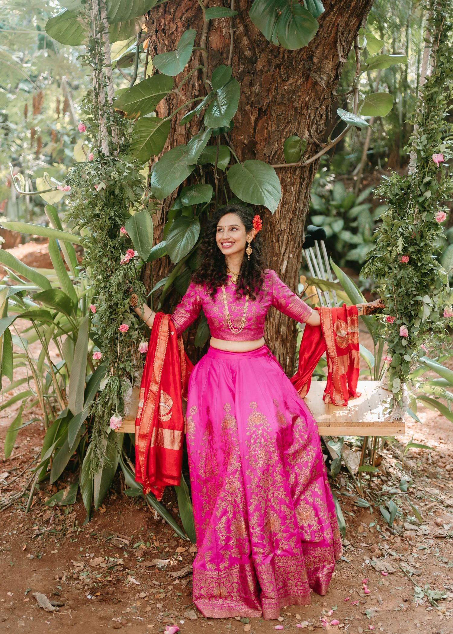 Woman in vibrant pink and gold lehenga with red dupatta seated on a swing surrounded by lush greenery and flowers.