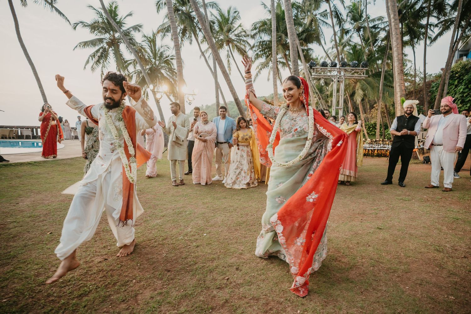 Couple dancing in traditional Indian wedding attire on a tropical lawn with palm trees and guests in background.