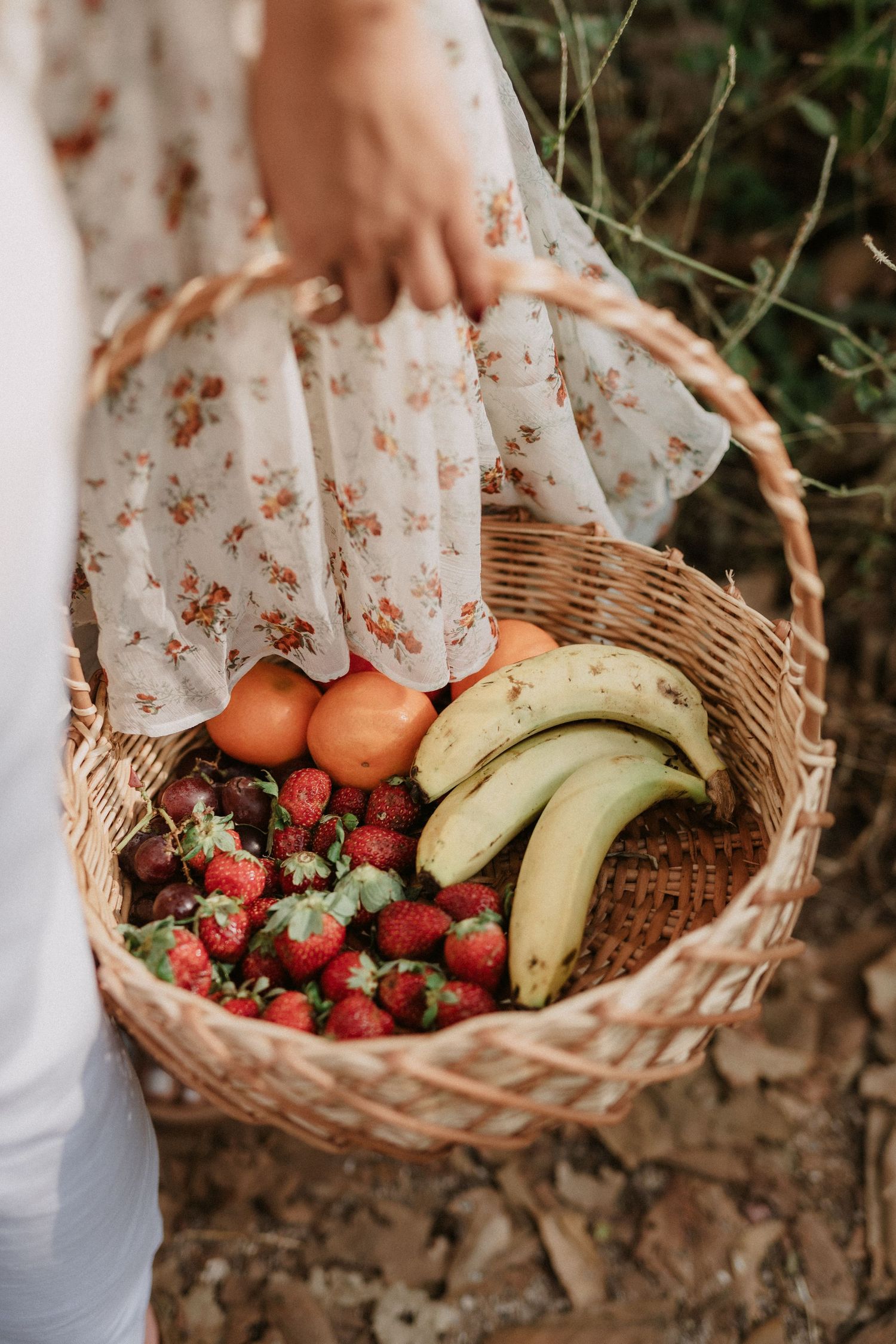 Person in floral dress holding wicker basket filled with fresh strawberries, bananas and oranges in garden setting.
