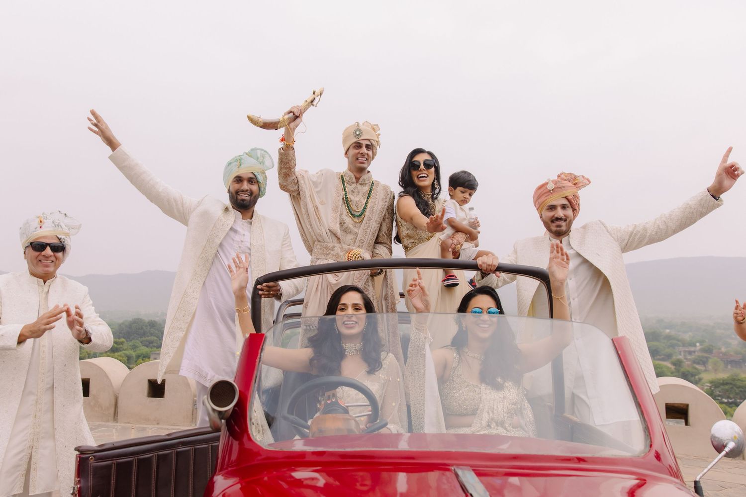 Wedding party celebrating in red convertible car with raised arms.