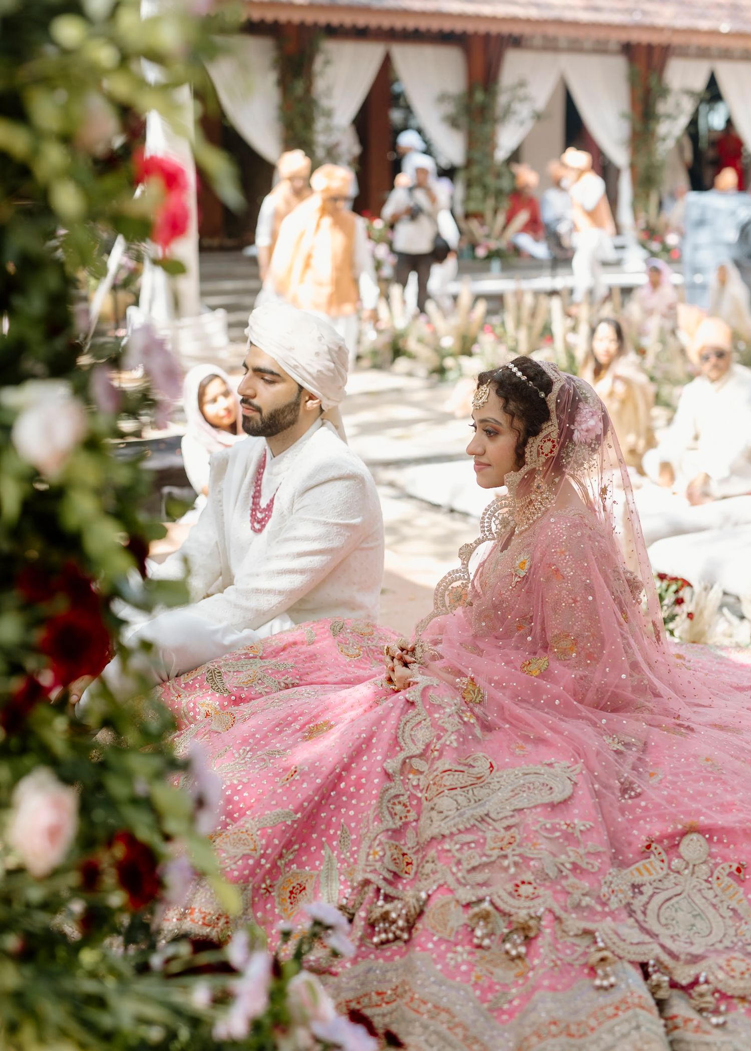 Traditional Indian wedding couple in pink and white attire sitting on ornate cushions during ceremony.