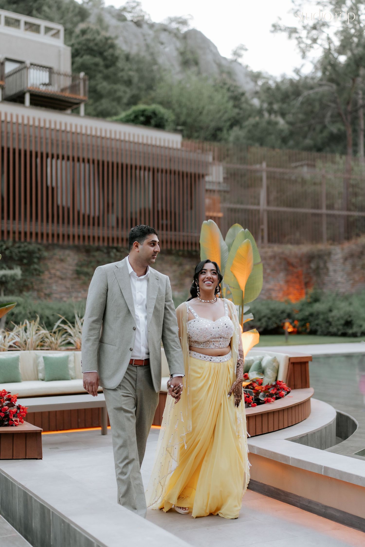 Indian couple in traditional attire walk hand-in-hand at an outdoor wedding venue with mountain backdrop.