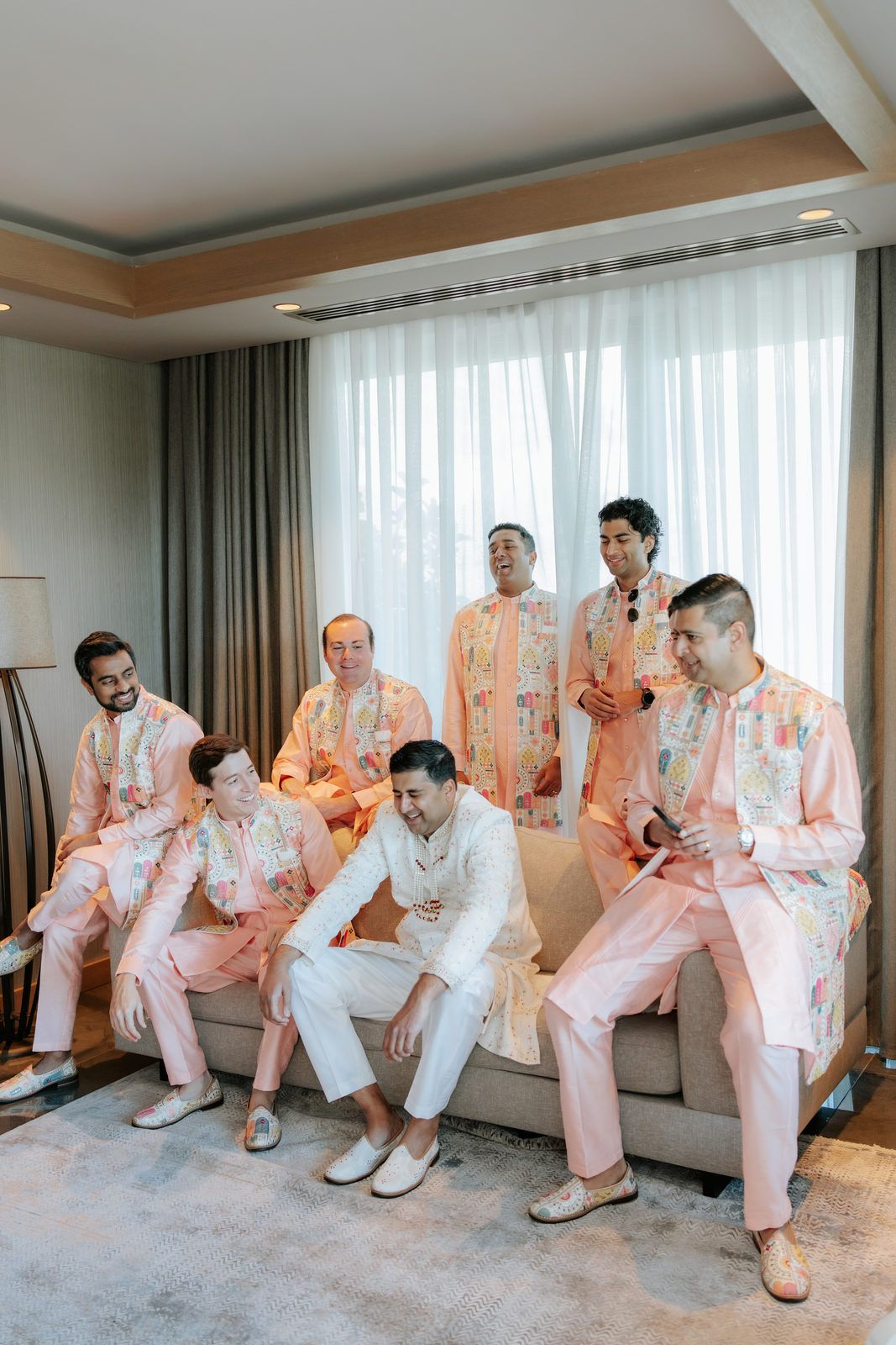 Groom in white sherwani poses with groomsmen in matching pink kurta pajamas in a hotel suite.