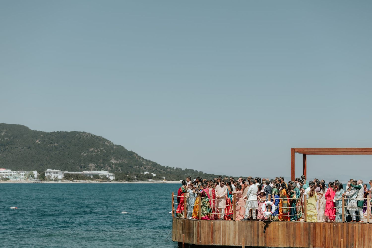 Guests gather on a wooden ocean pier for a wedding ceremony with turquoise water and green hills backdrop.