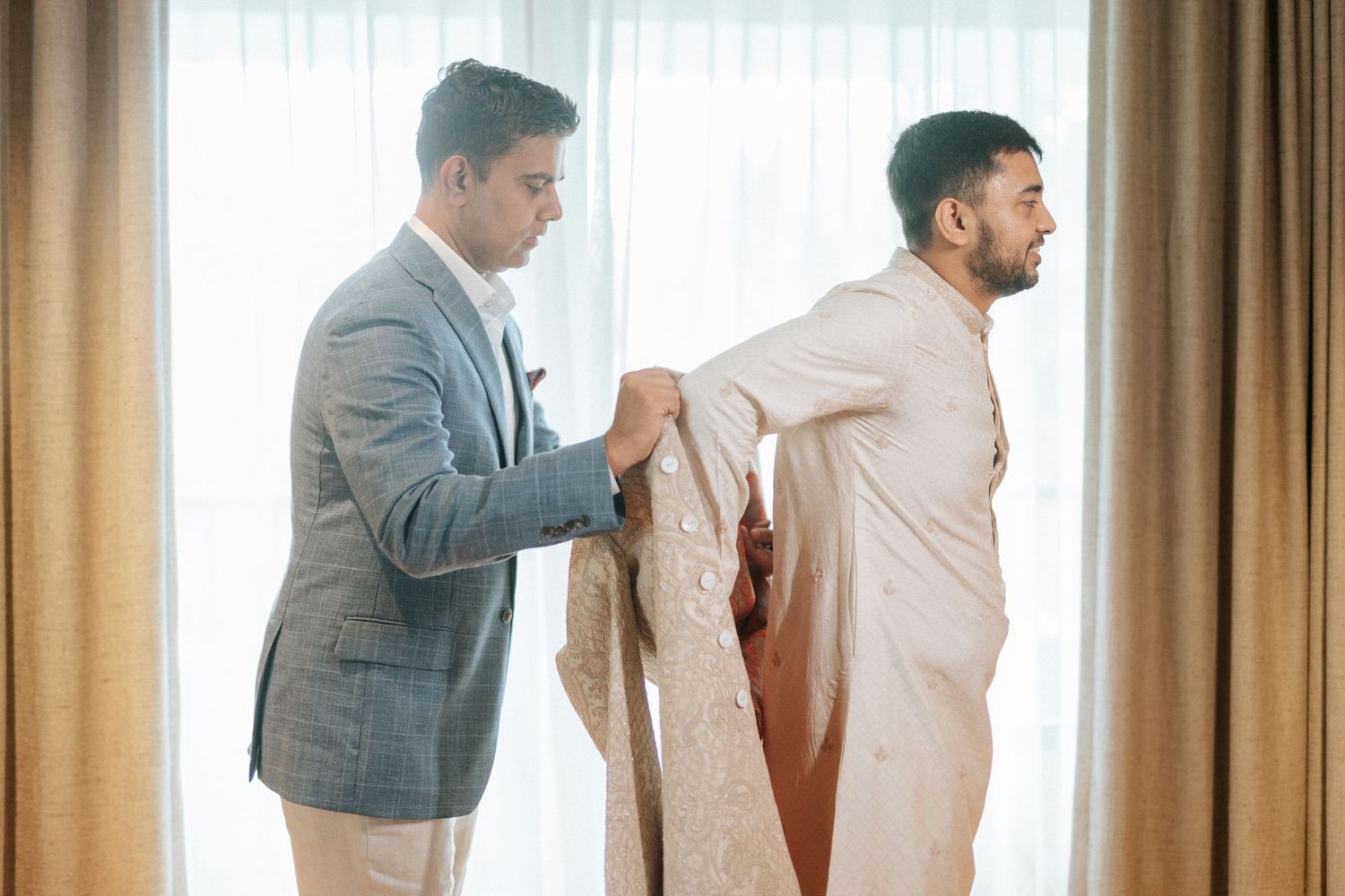 A man in a gray suit helps another man dress in traditional South Asian wedding attire near a bright window.