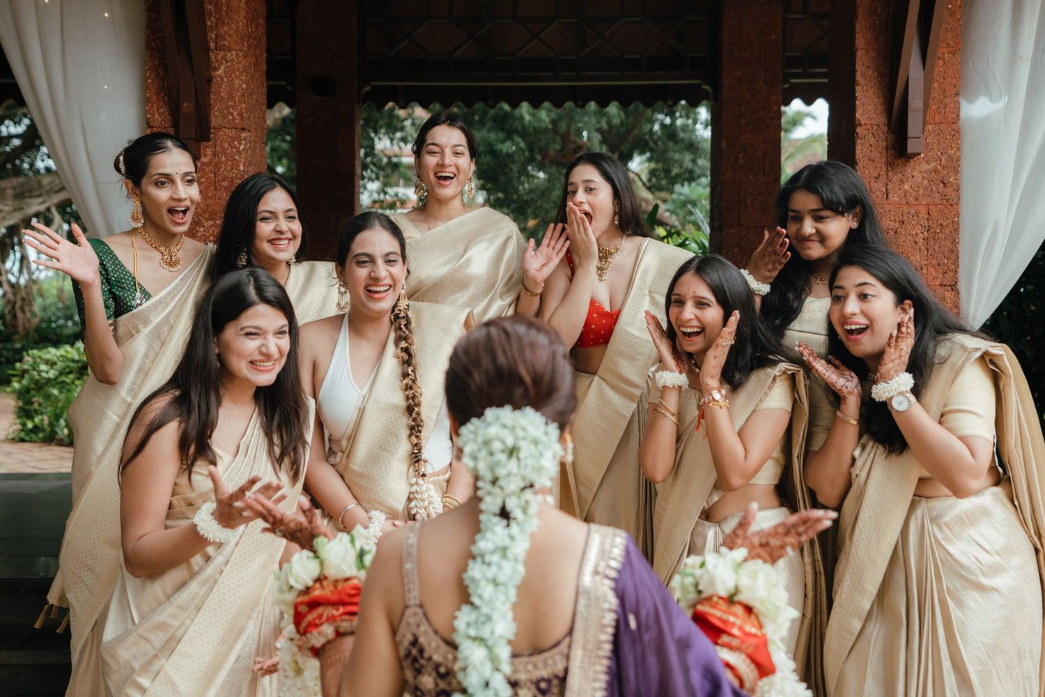 Bridesmaids in gold sarees react with joy and surprise to a bride holding flowers at an Indian wedding celebration.