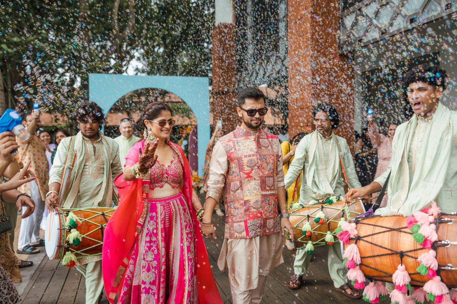 Vibrant Indian wedding procession with couple in pink attire, dhol drummers, and flower confetti celebration.