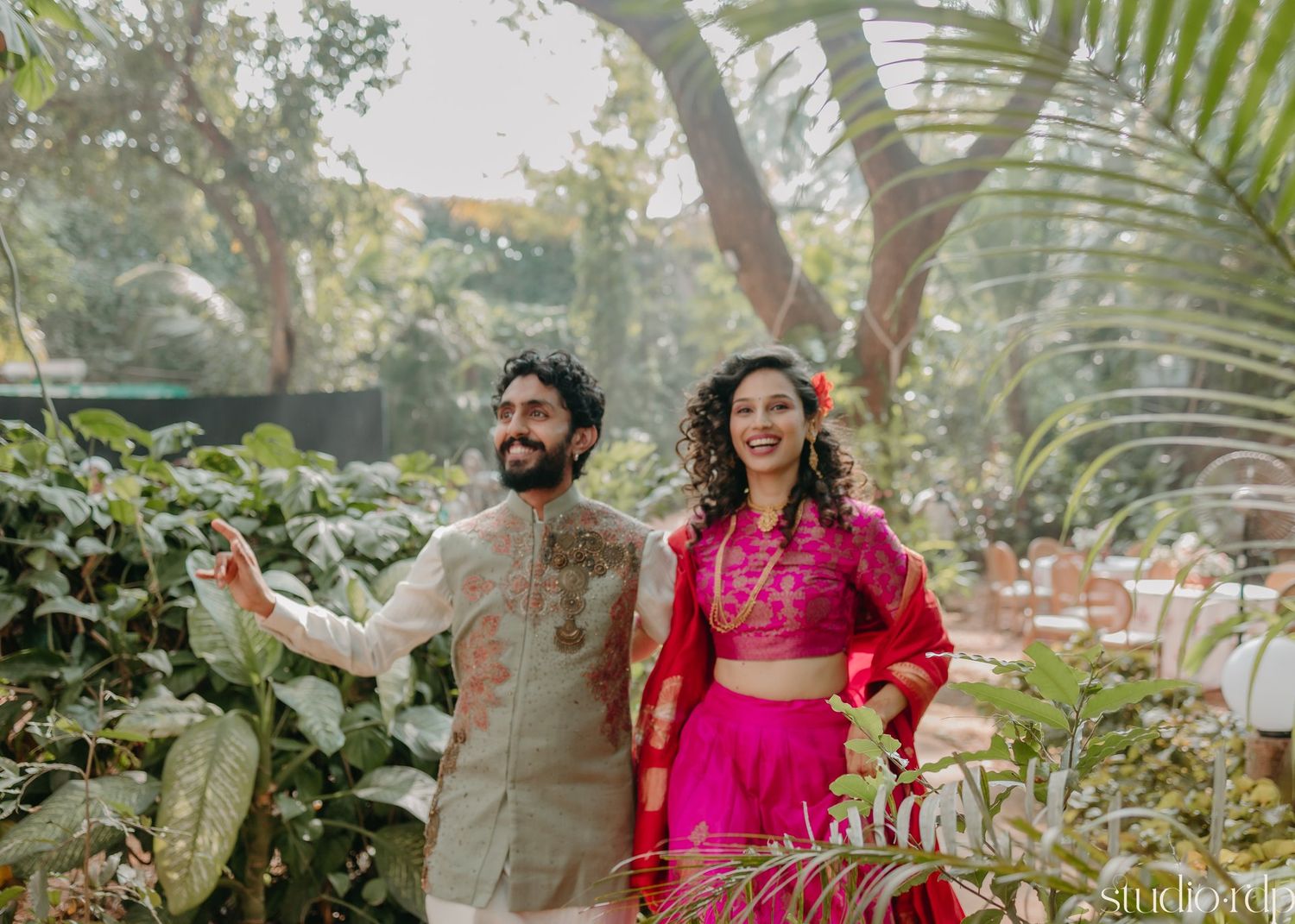 Couple in traditional Indian attire posing in a lush garden setting with palm trees.