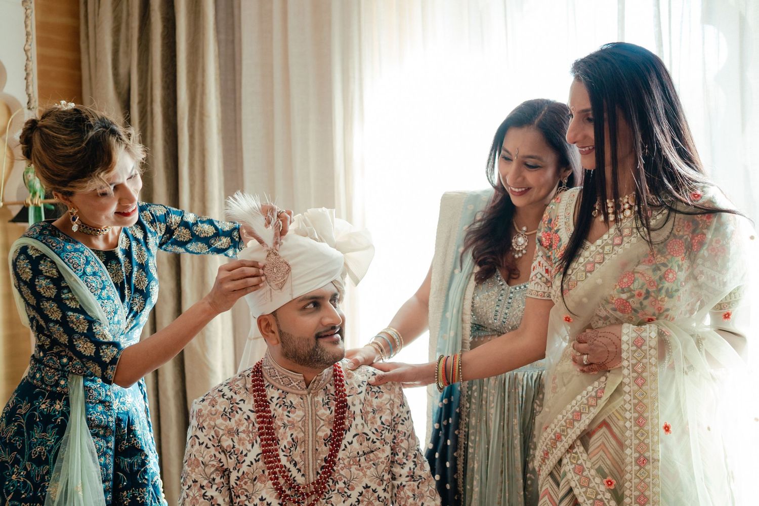 A groom in traditional Indian attire sits while family members help prepare him for a wedding ceremony.