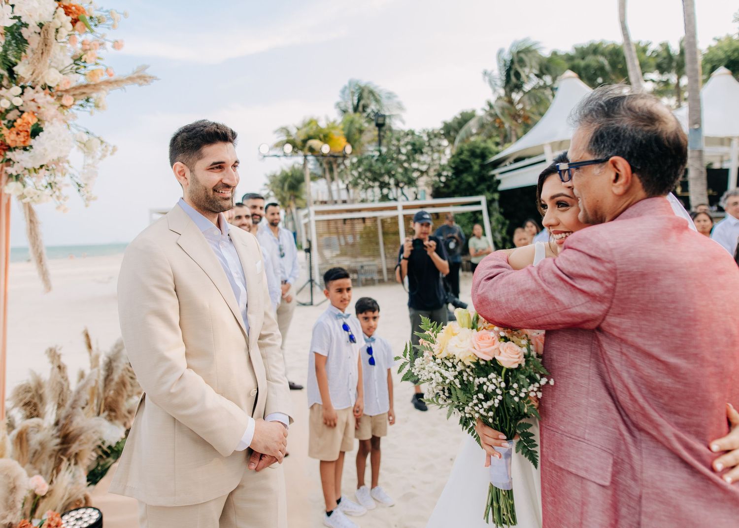 A beachside wedding ceremony setup with attendees in neutral-colored attire amongst palm trees and pampas grass decor.