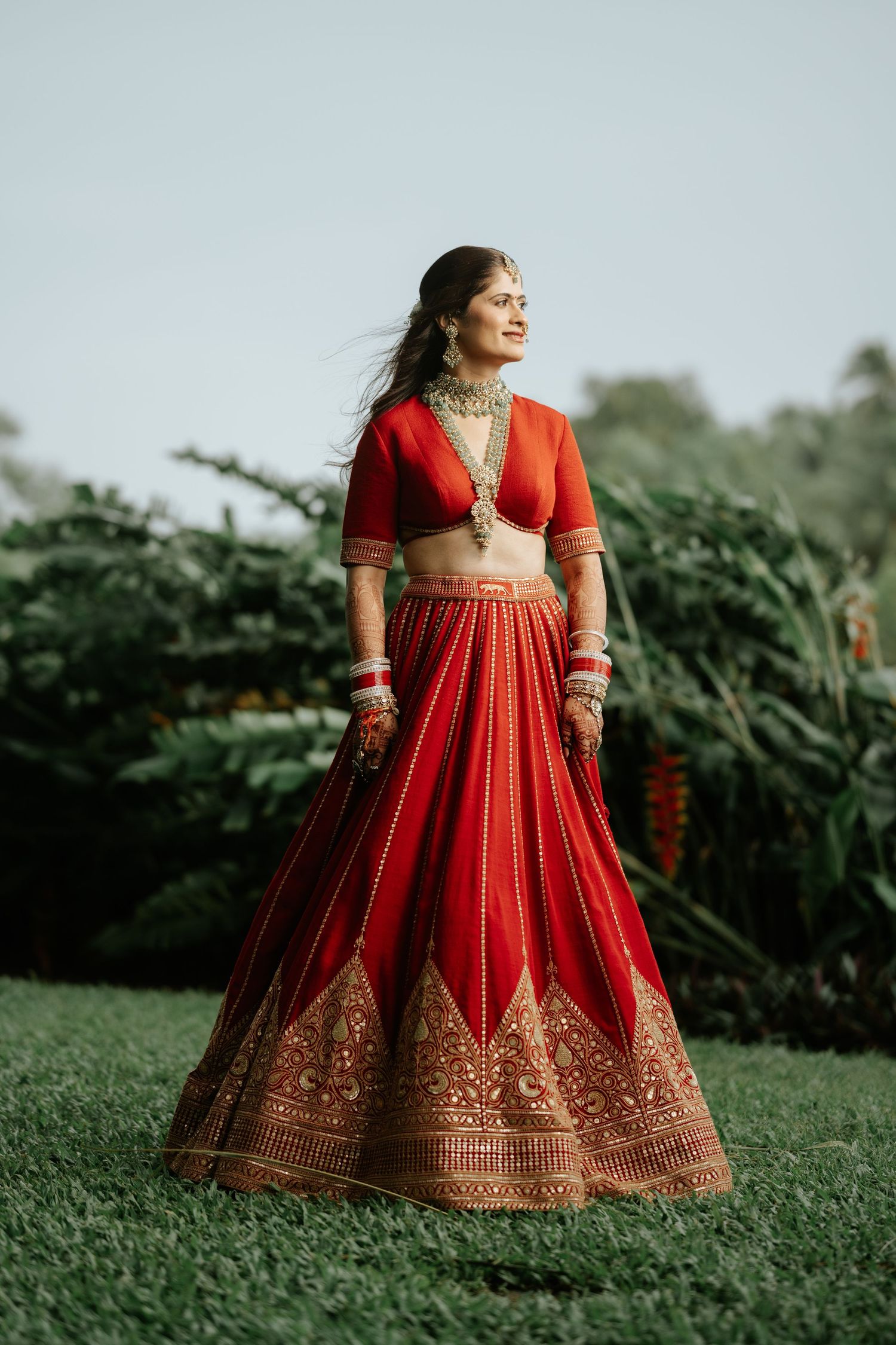 Bride in red embroidered lehenga choli with gold jewelry poses outdoors in lush garden setting.