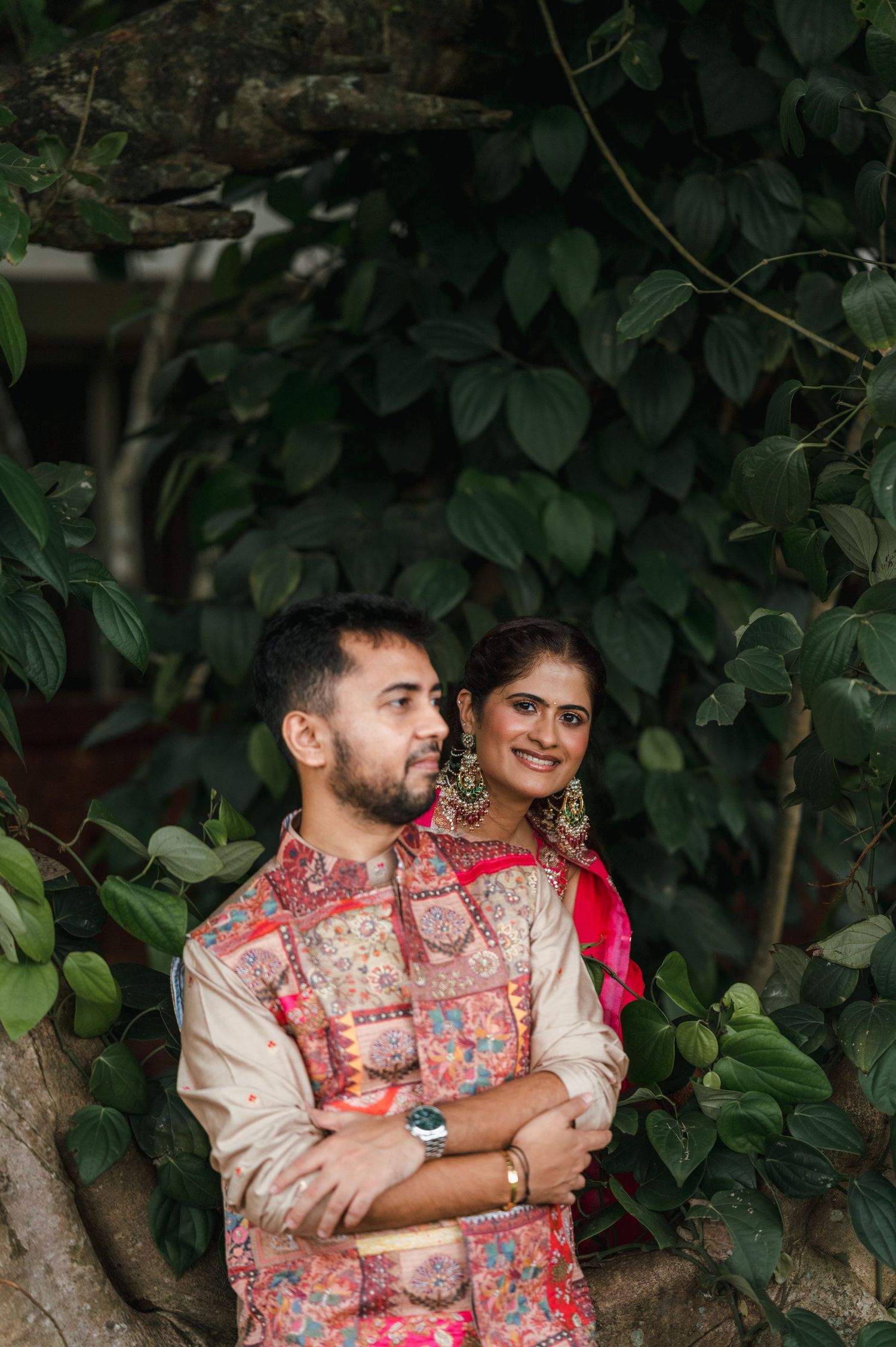 Indian couple in traditional attire embracing outdoors, surrounded by lush green foliage, smiling joyfully.