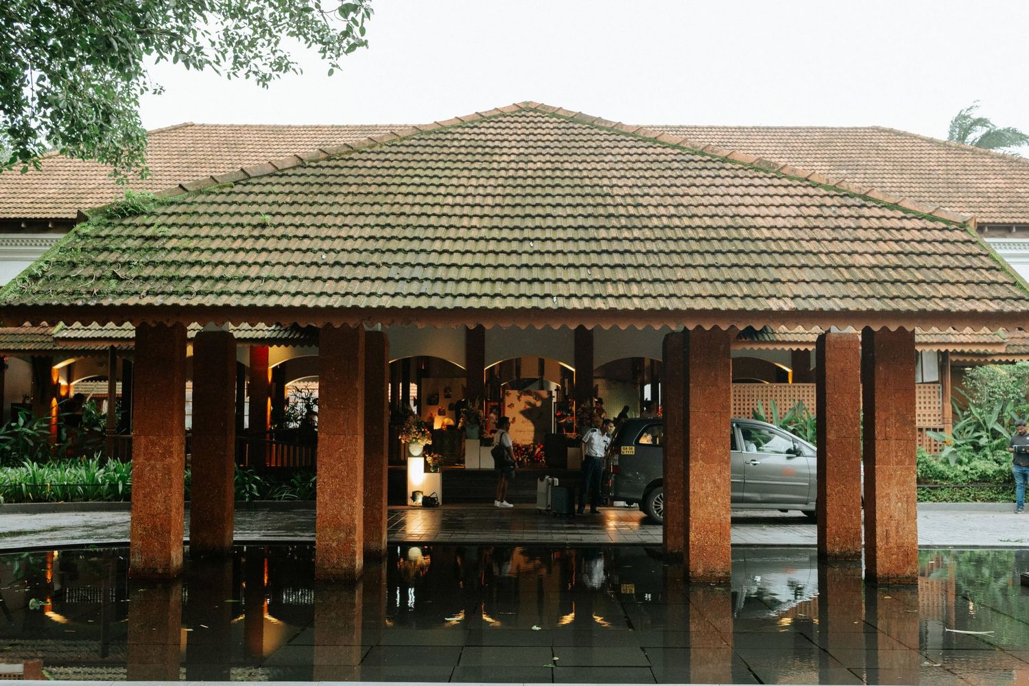 Open-air tropical pavilion with terracotta tile roof, wooden pillars, and reflective wet floors surrounded by lush greenery.