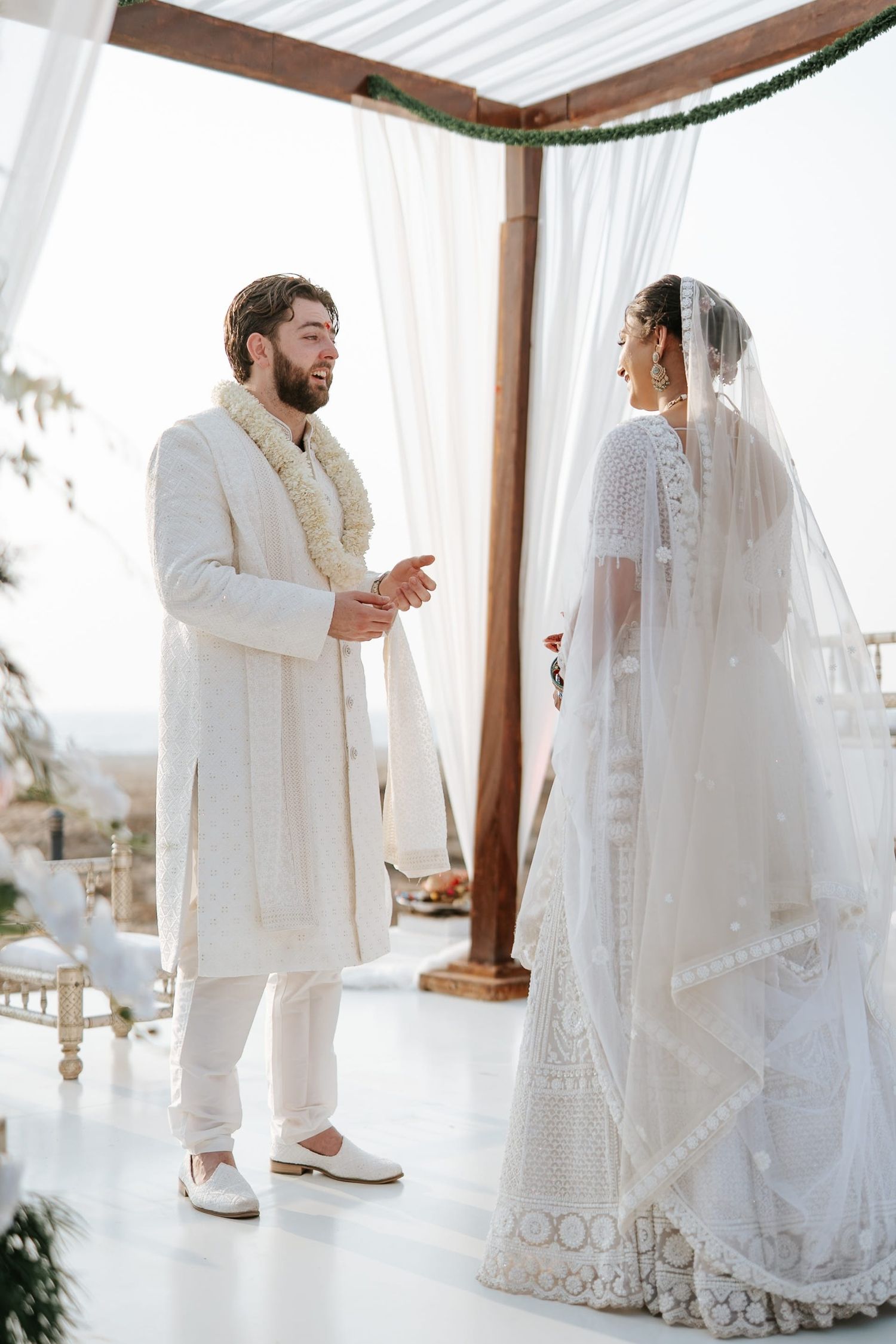 Bride and groom in white traditional Indian wedding attire exchange vows under a wooden mandap outdoors.