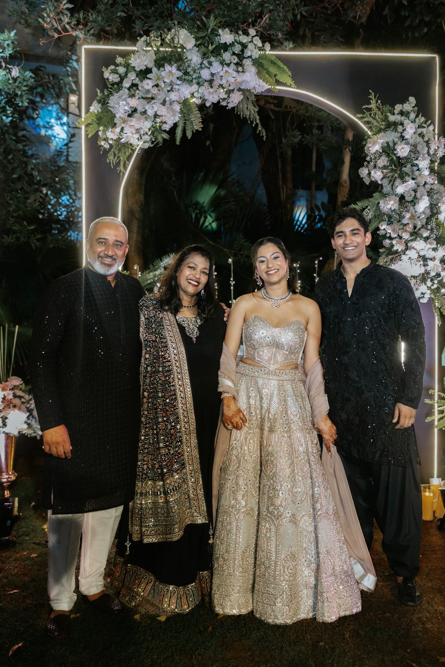 Family of four posing under floral arch at Indian wedding reception, wearing elegant traditional outfits.